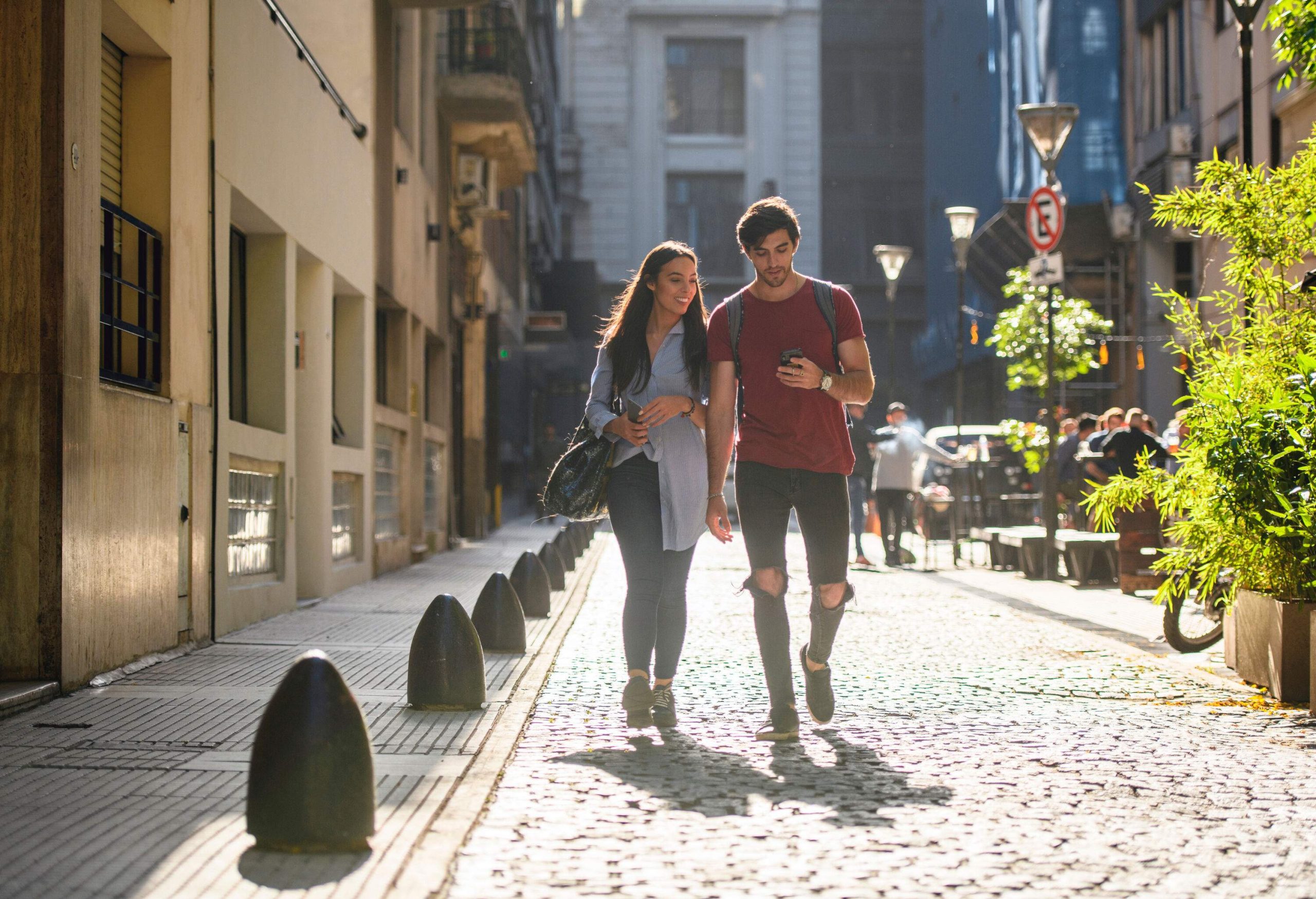 A couple looks at the man's smartphone while walking on the cobbled street enclosed by tall buildings.