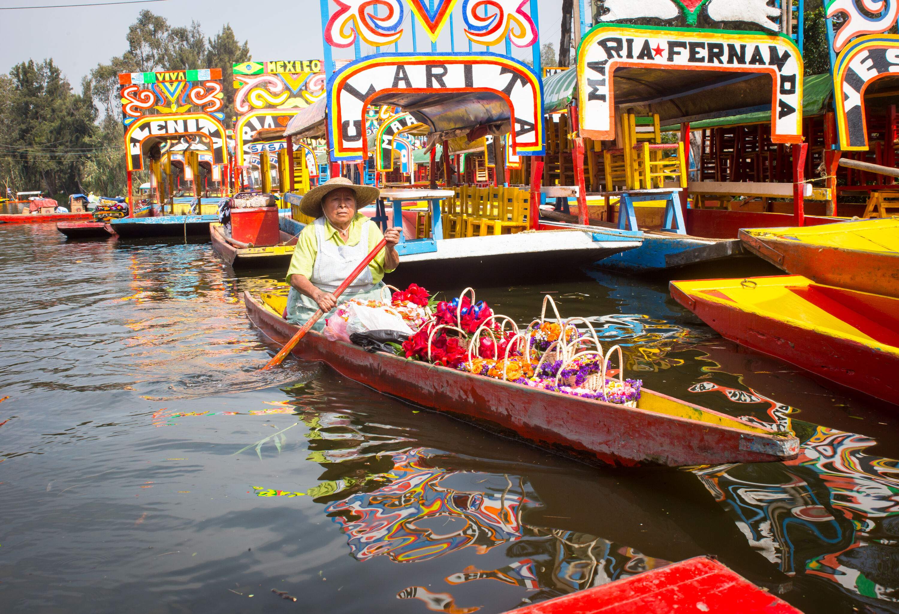 A woman in a row boat carrying multicoloured flower baskets passing by the trajineras floating in the water.