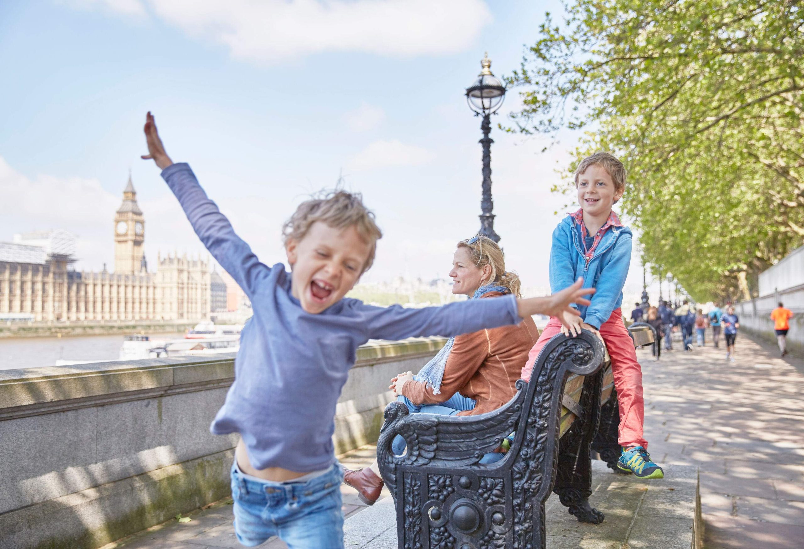 A boy runs with his arms spread out in front of a woman and a child sitting on a sidewalk bench.