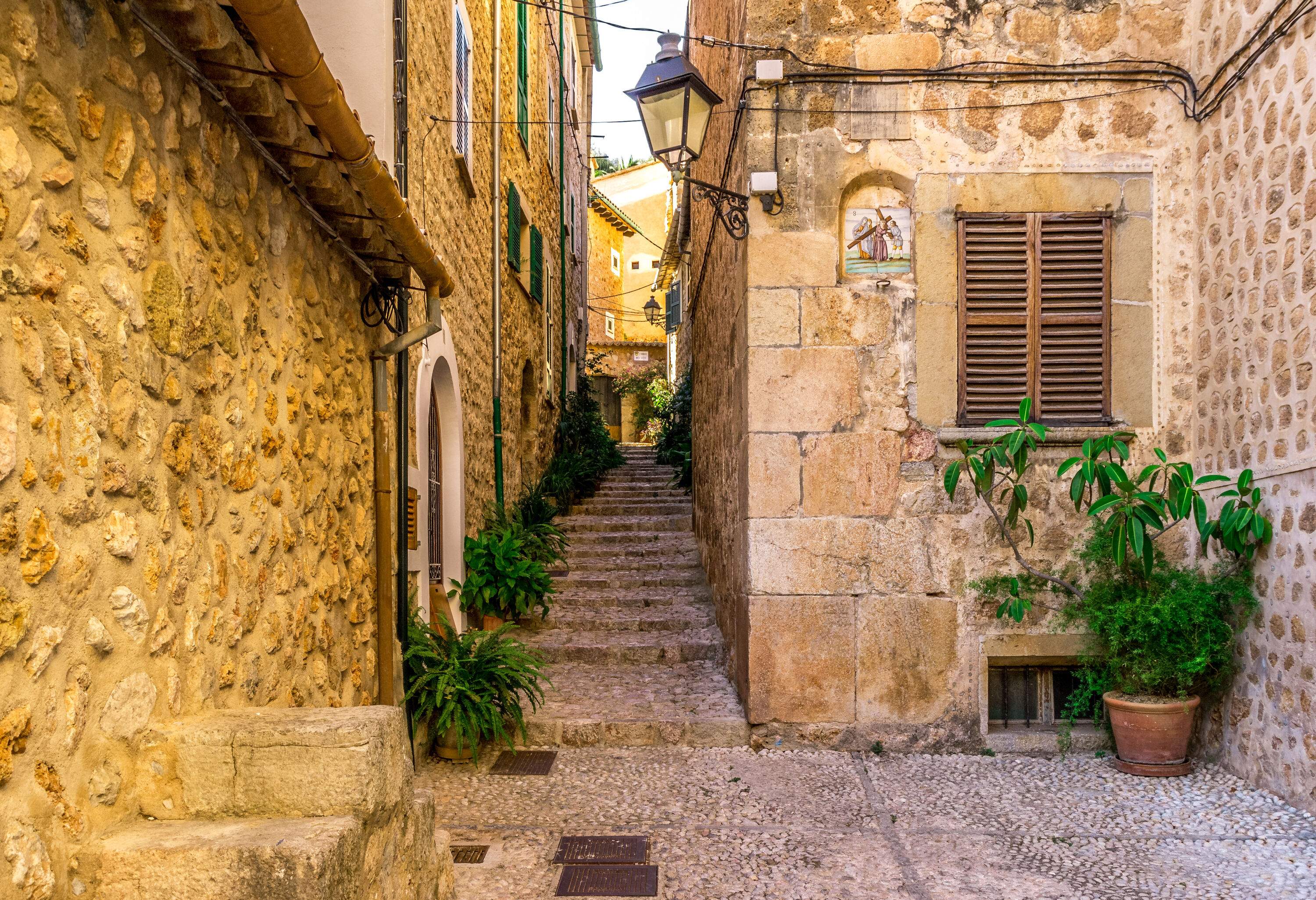 A cobblestone-paved alley with steps between stone homes and plant pots on the side.