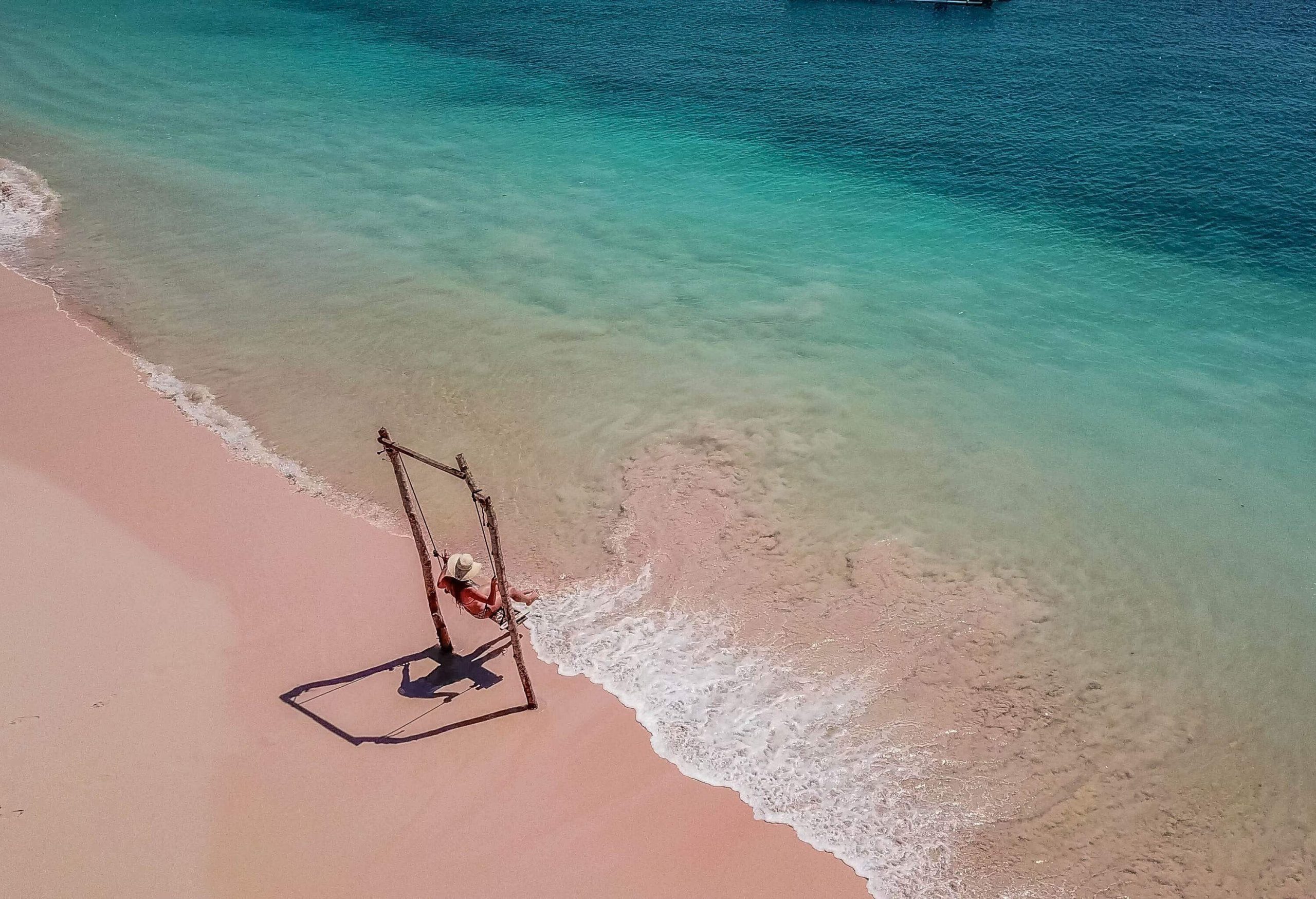 A girl in a wide-brimmed hat sits on a beach swing set on a pink sand beach.