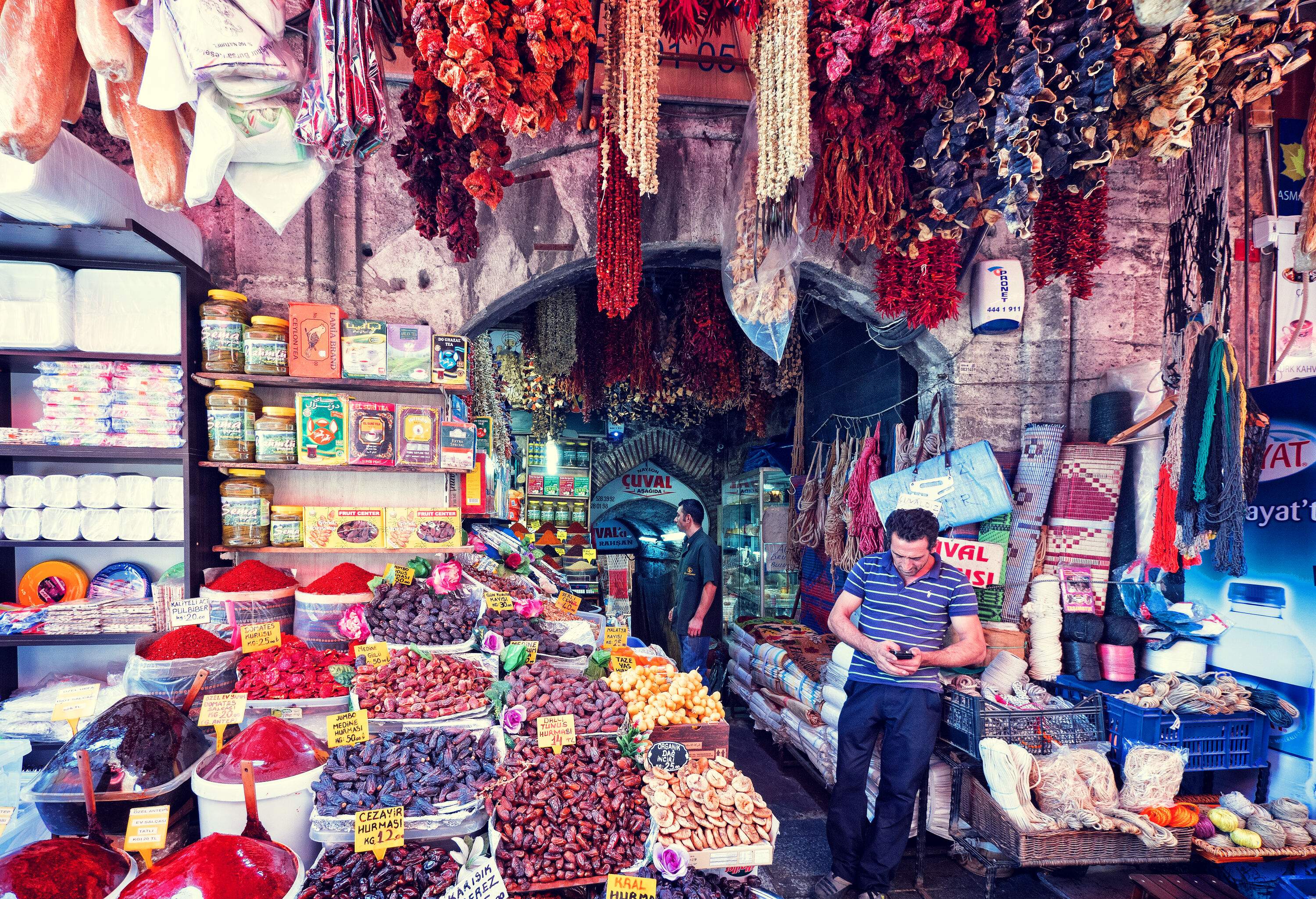 A man stands at the entrance of a bazaar with a display of assorted spices, carpets, and souvenir items.