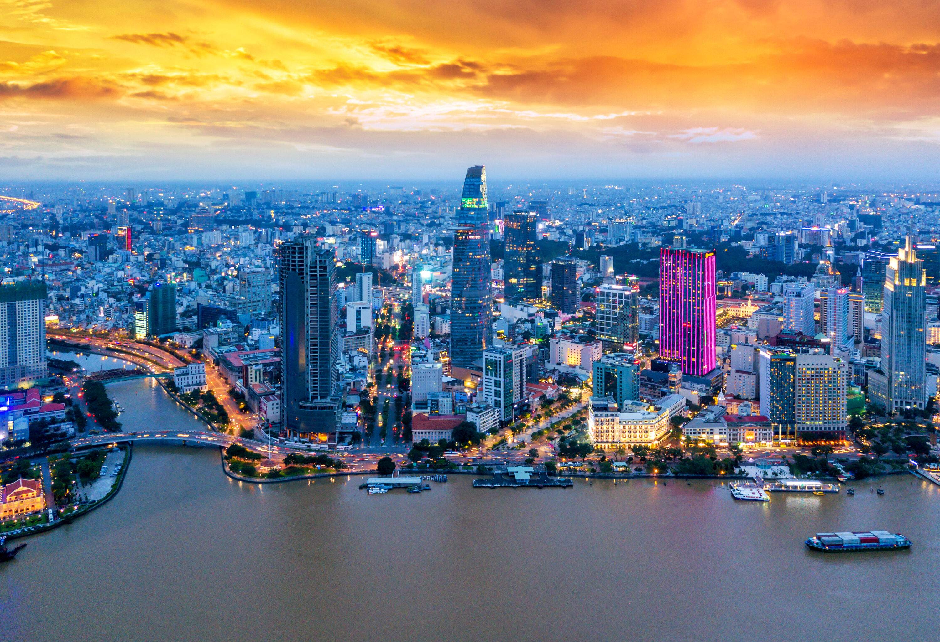 Aerial view of a brightly lit city with modern towering buildings by the river under the scenic orangy twilight sky.