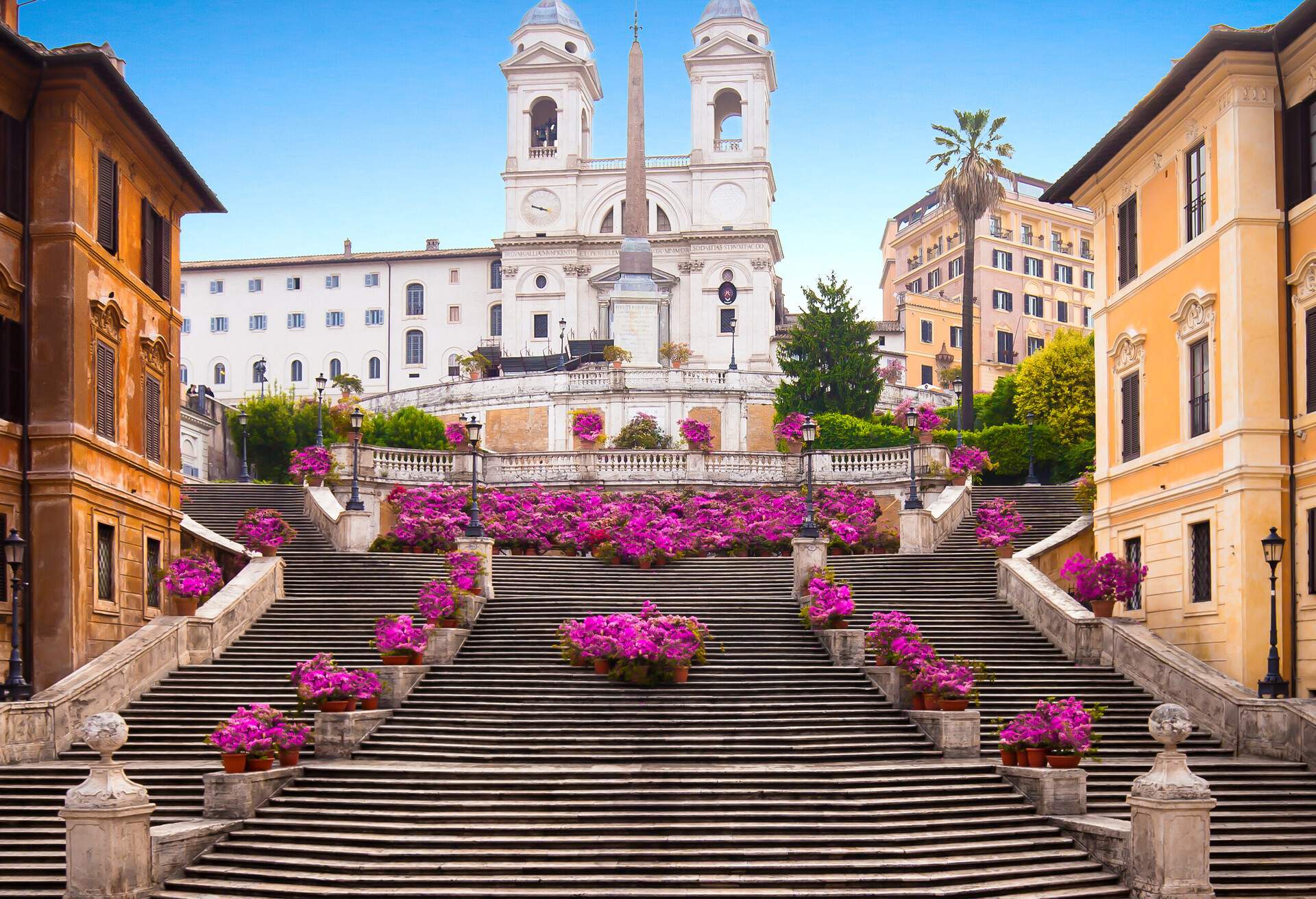 A Spanish steps adorned with purple flowering plants surrounded by classic buildings towards a renaissance church with two bell towers.