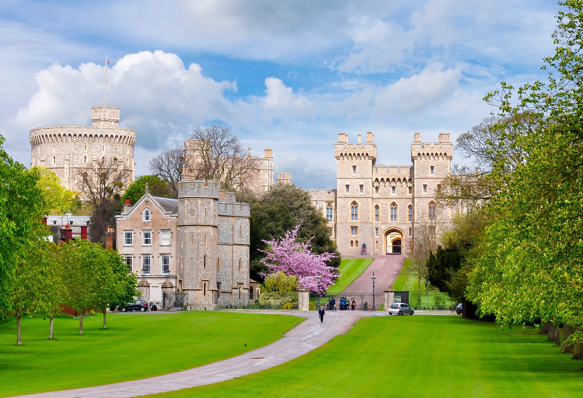 A gate to a castle at the end of a road, beyond an iron fence and surrounded by verdant lawns and trees.