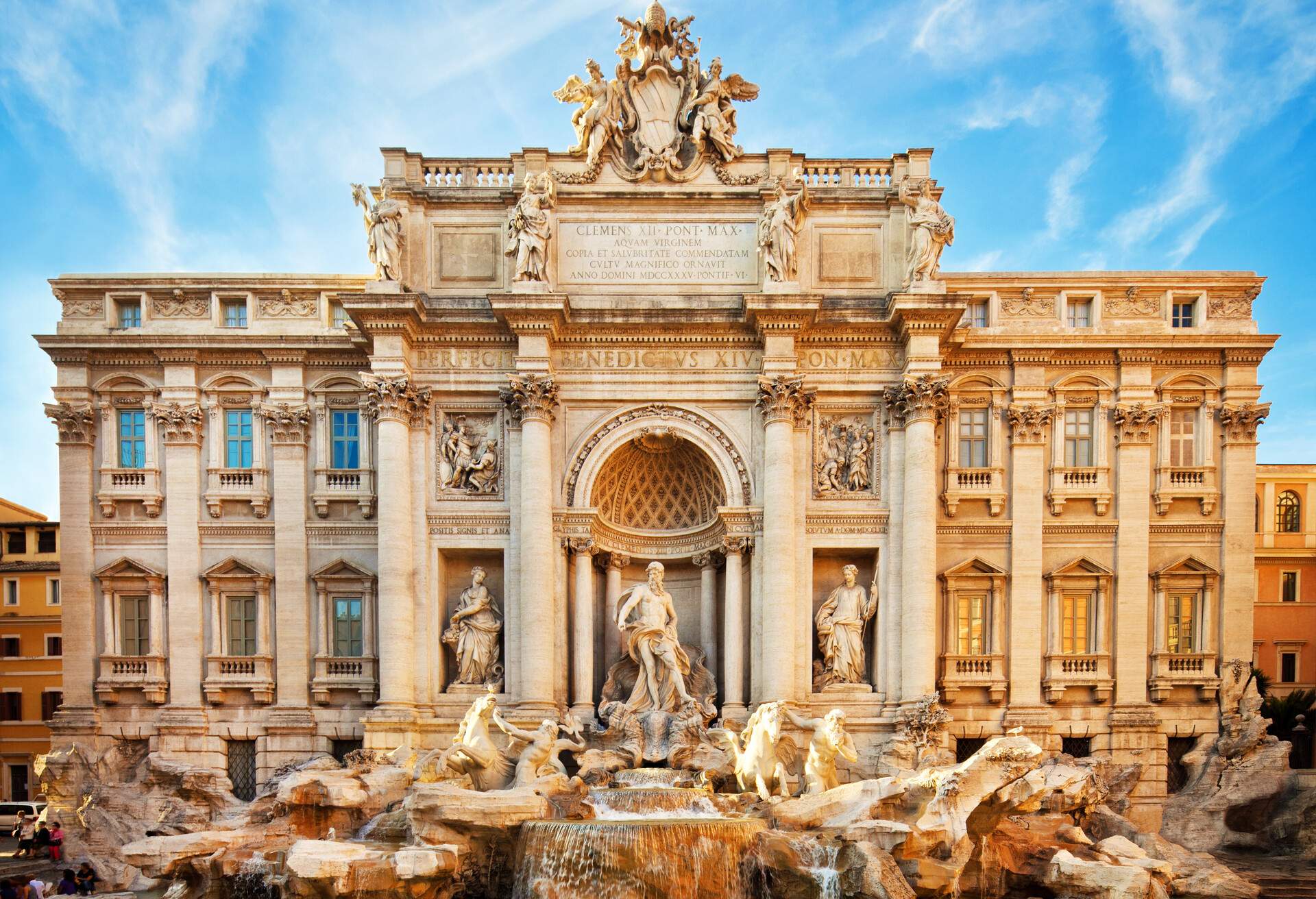 A medieval Baroque fountain with a backdrop of a palace with three statues on the free-standing columns and water falling on the rockwork.