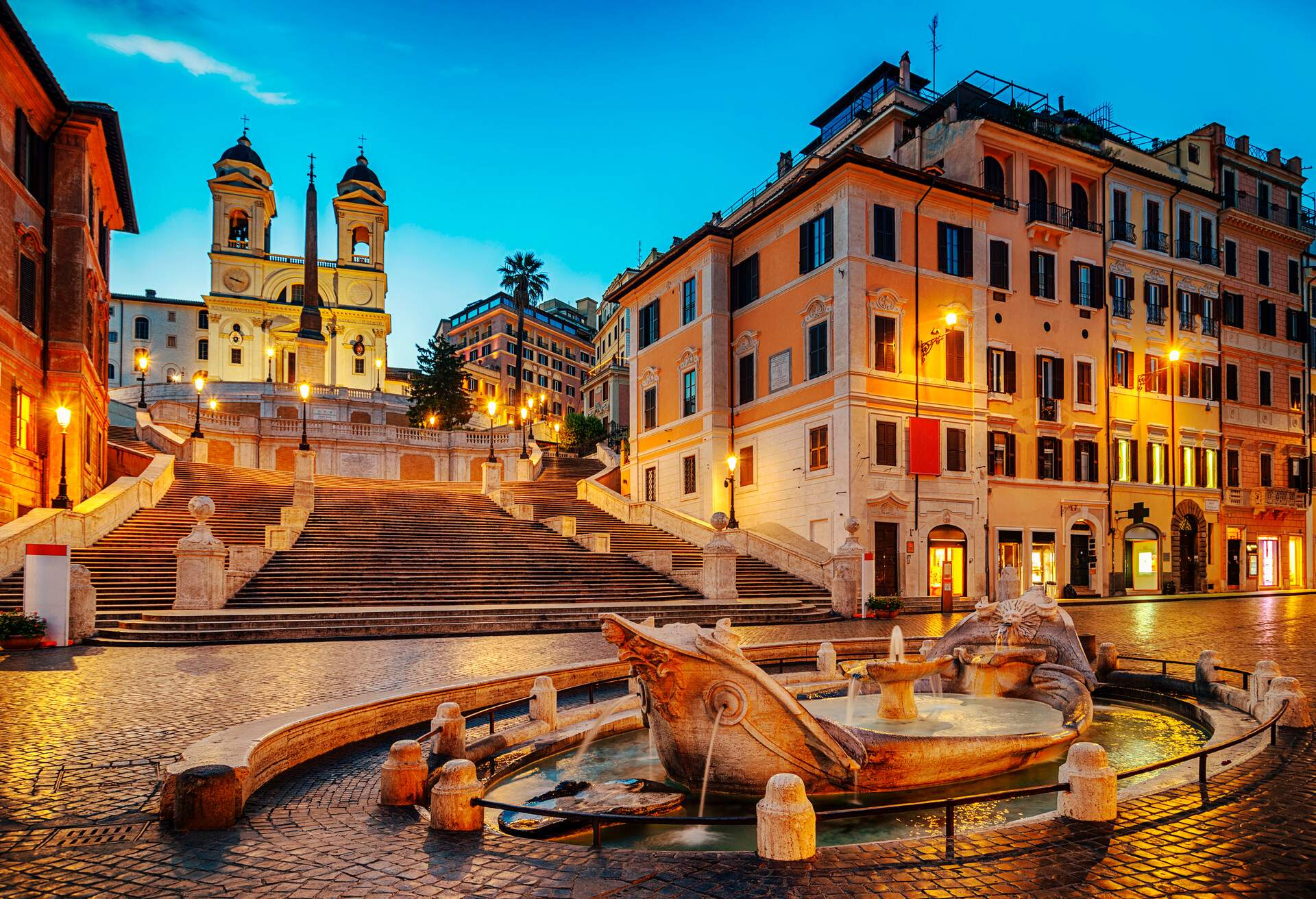 A boat fountain at the foot of an illuminated stairway in a cobbled square.