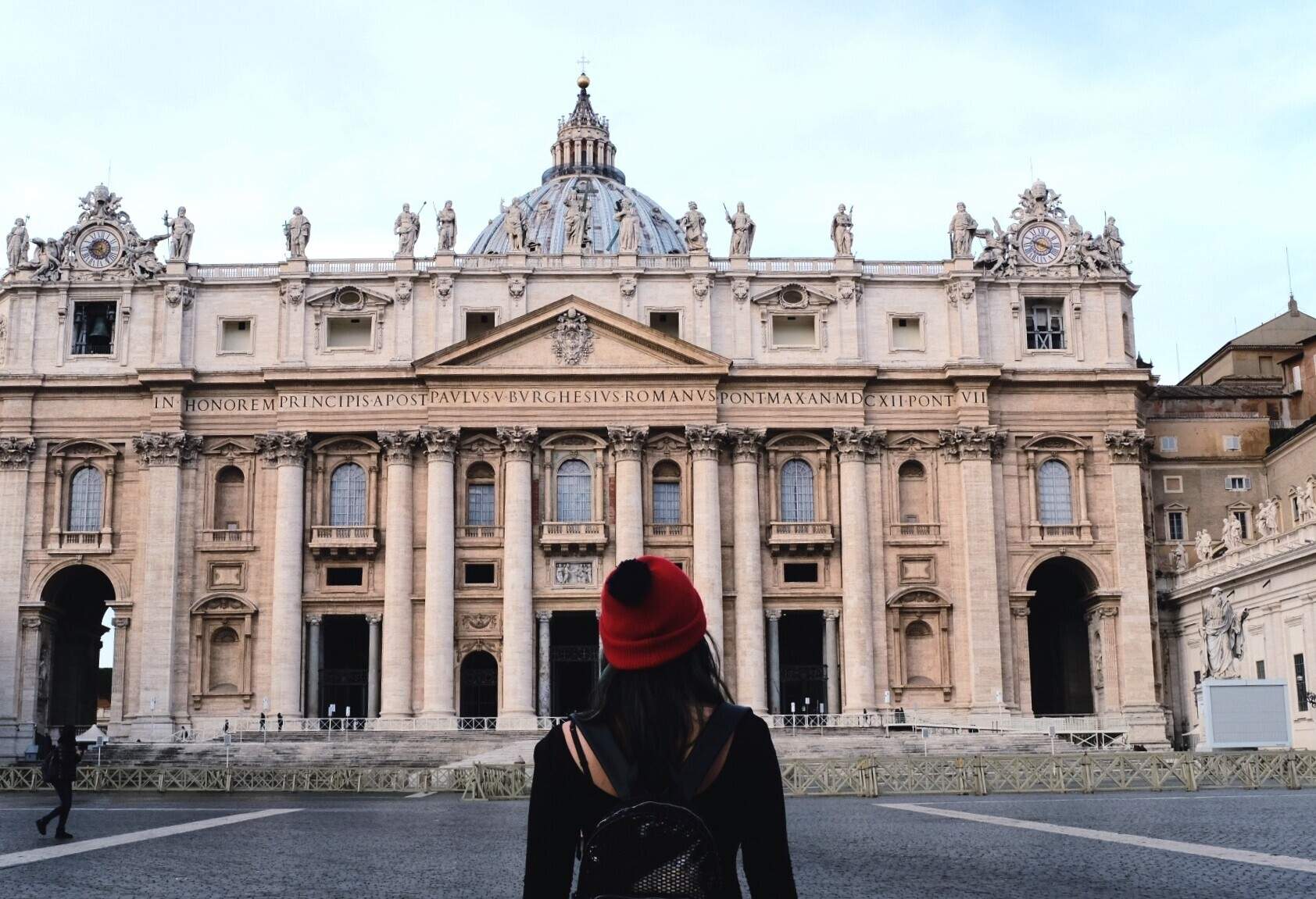 An individual in front of the main facade of the domed, Late Renaissance-style St. Peter's Basilica in Vatican City.