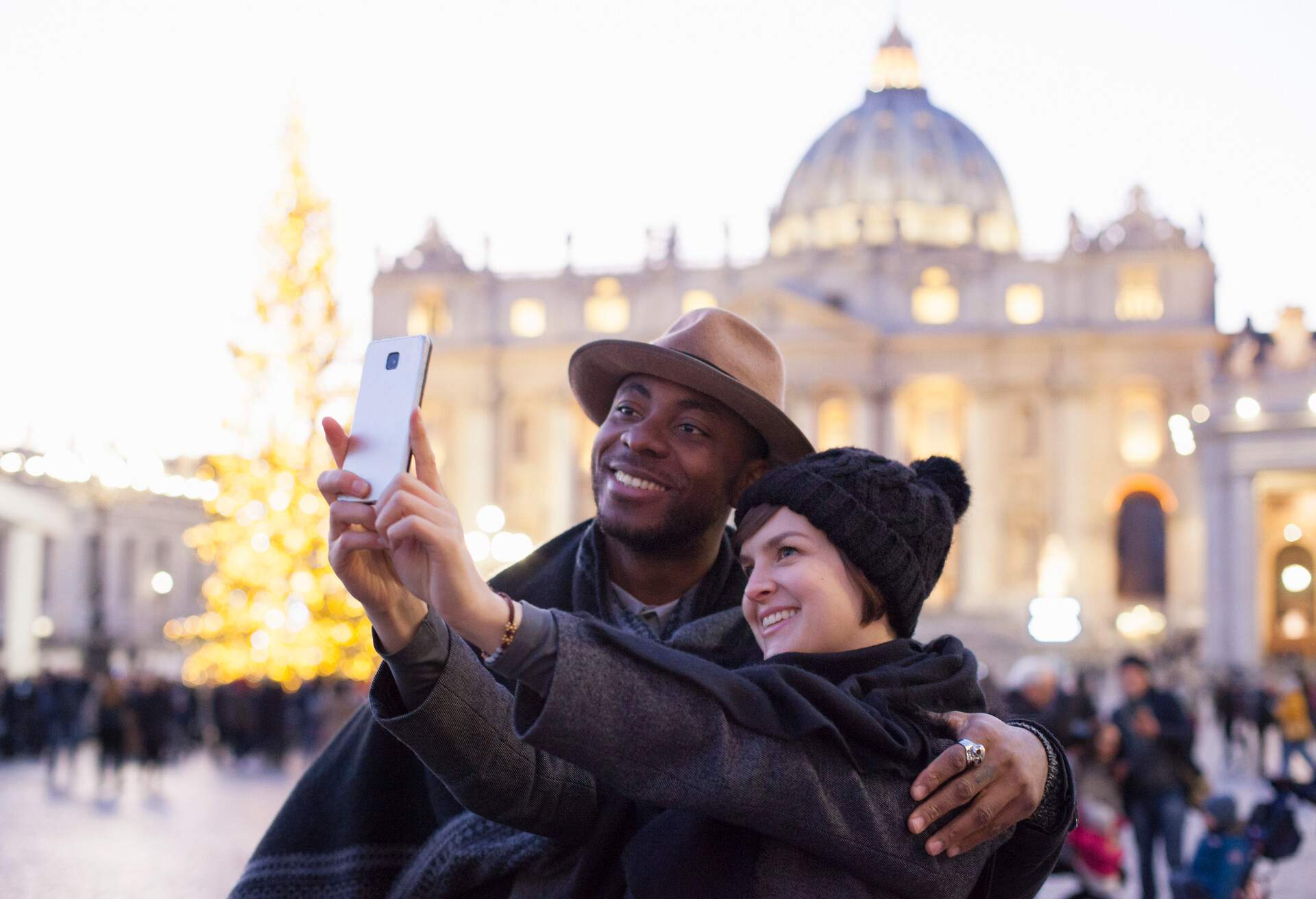 A man and a woman taking a picture together on a plaza with a Renaissance style church and a lit up Christmas tree in the background.