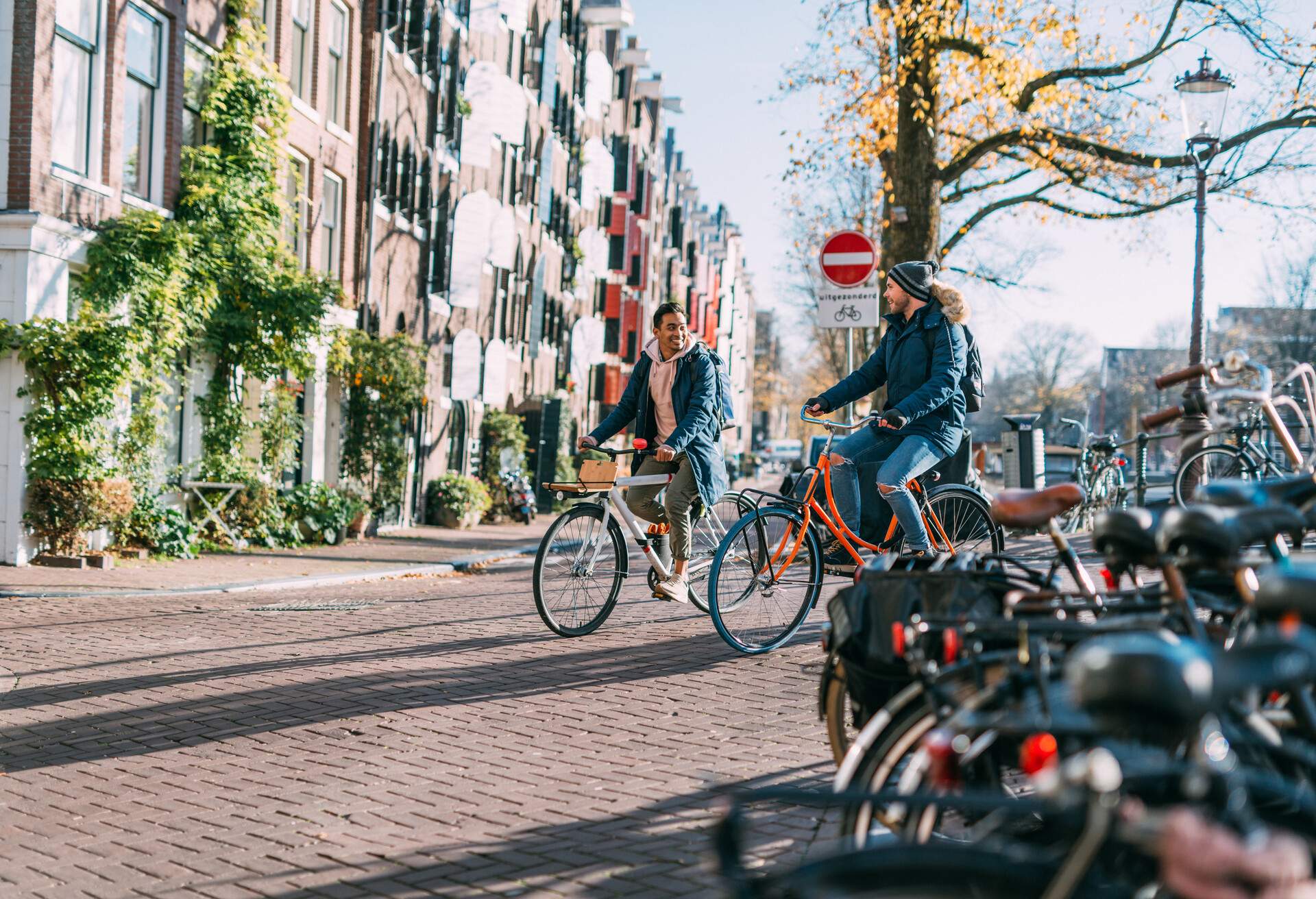 A happy couple relishes their city break as they ride bicycles along a sidewalk, passing by a charming row of buildings and parked bicycles.