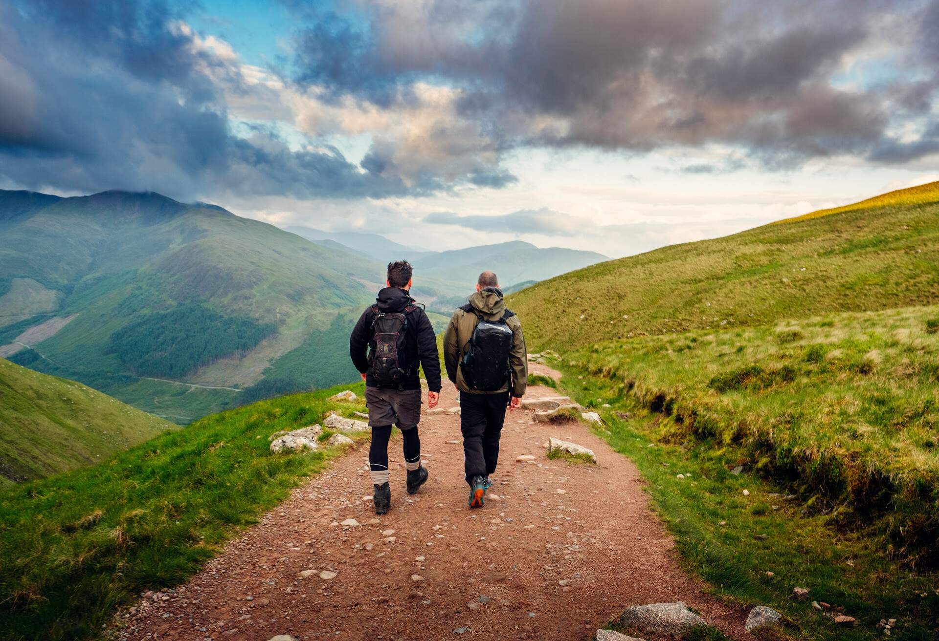 Two men walk along a sandy trail looking at a green mountain landscape and a cloudy dark sky.