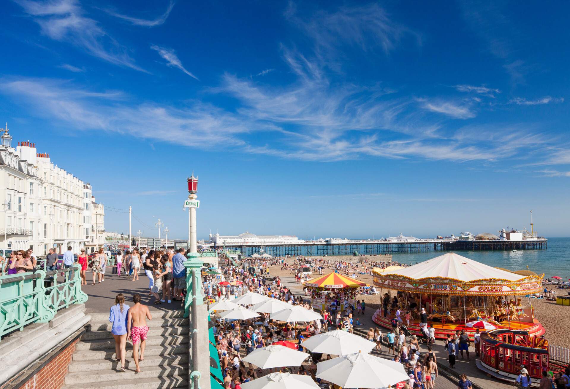 A seafront carnival crowded with people along a carousel, tent shops, arcades, and rides.