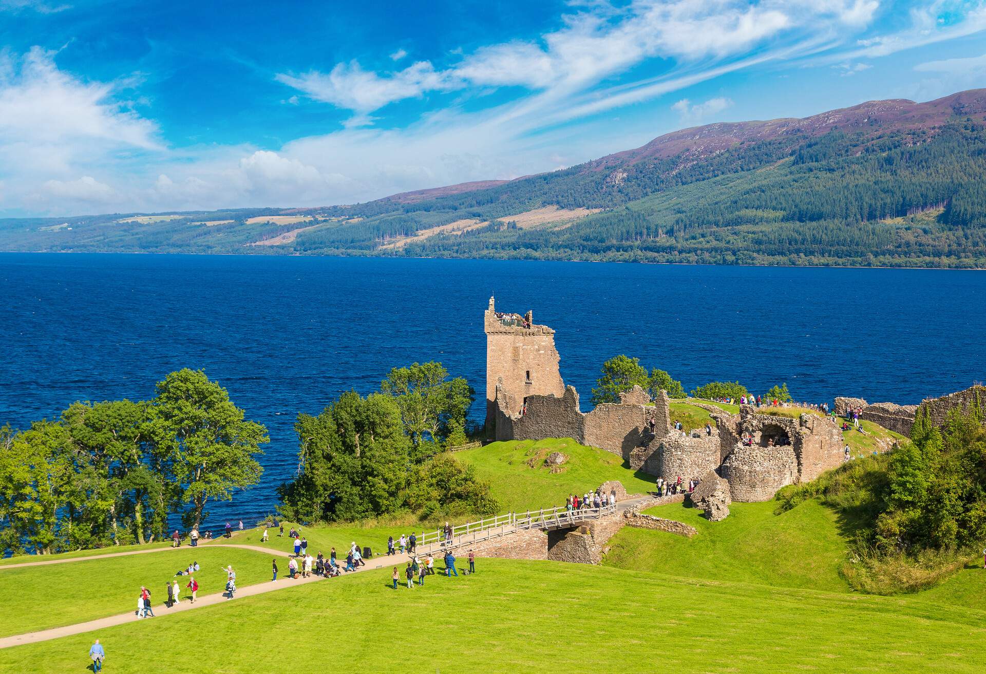 Crumbling stone structure near Loch Ness lake that is frequented by visitors may be seen in the background of a long mountain slope.