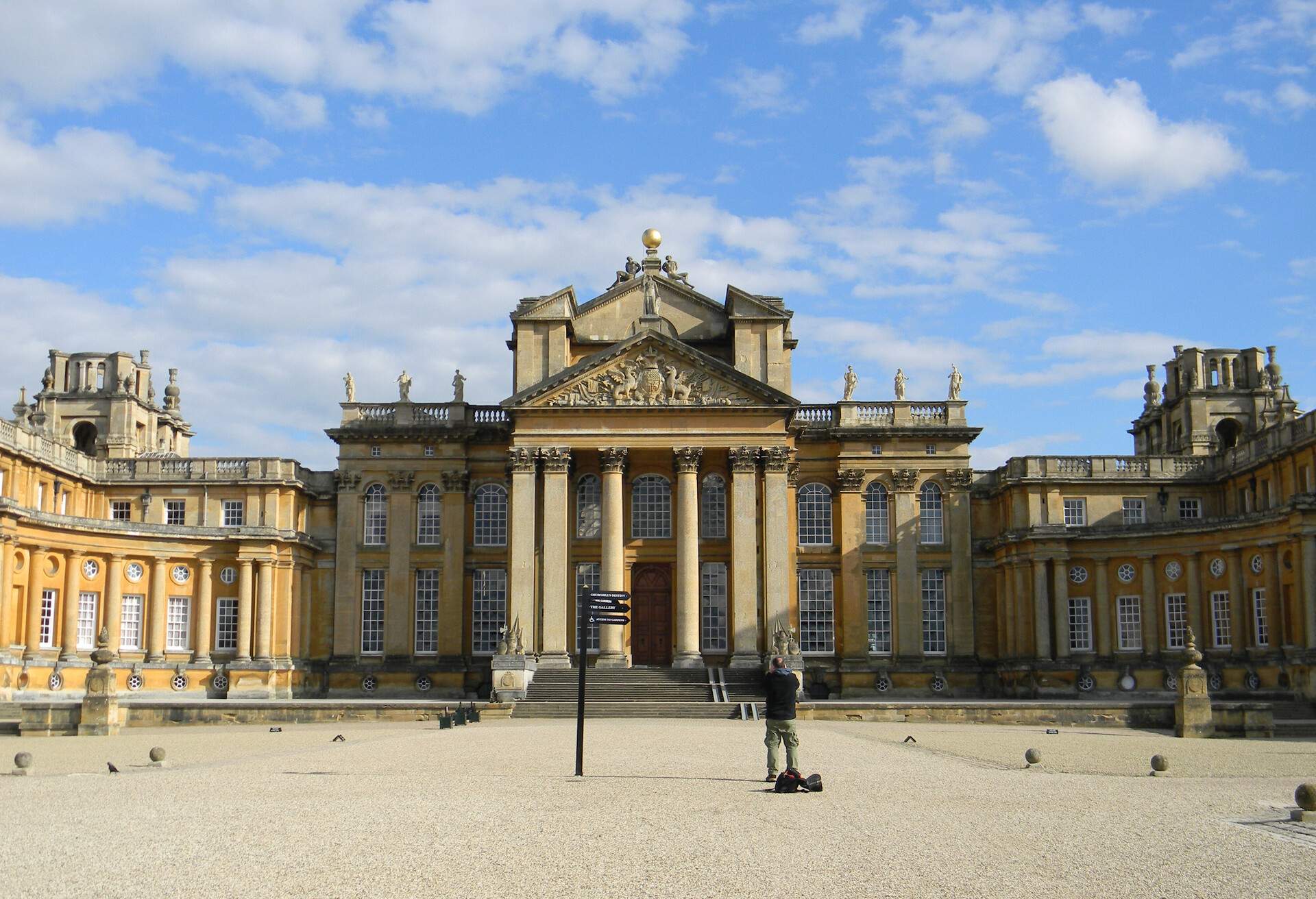 A person standing on gravel is photographing the huge, baroque-style English palace.
