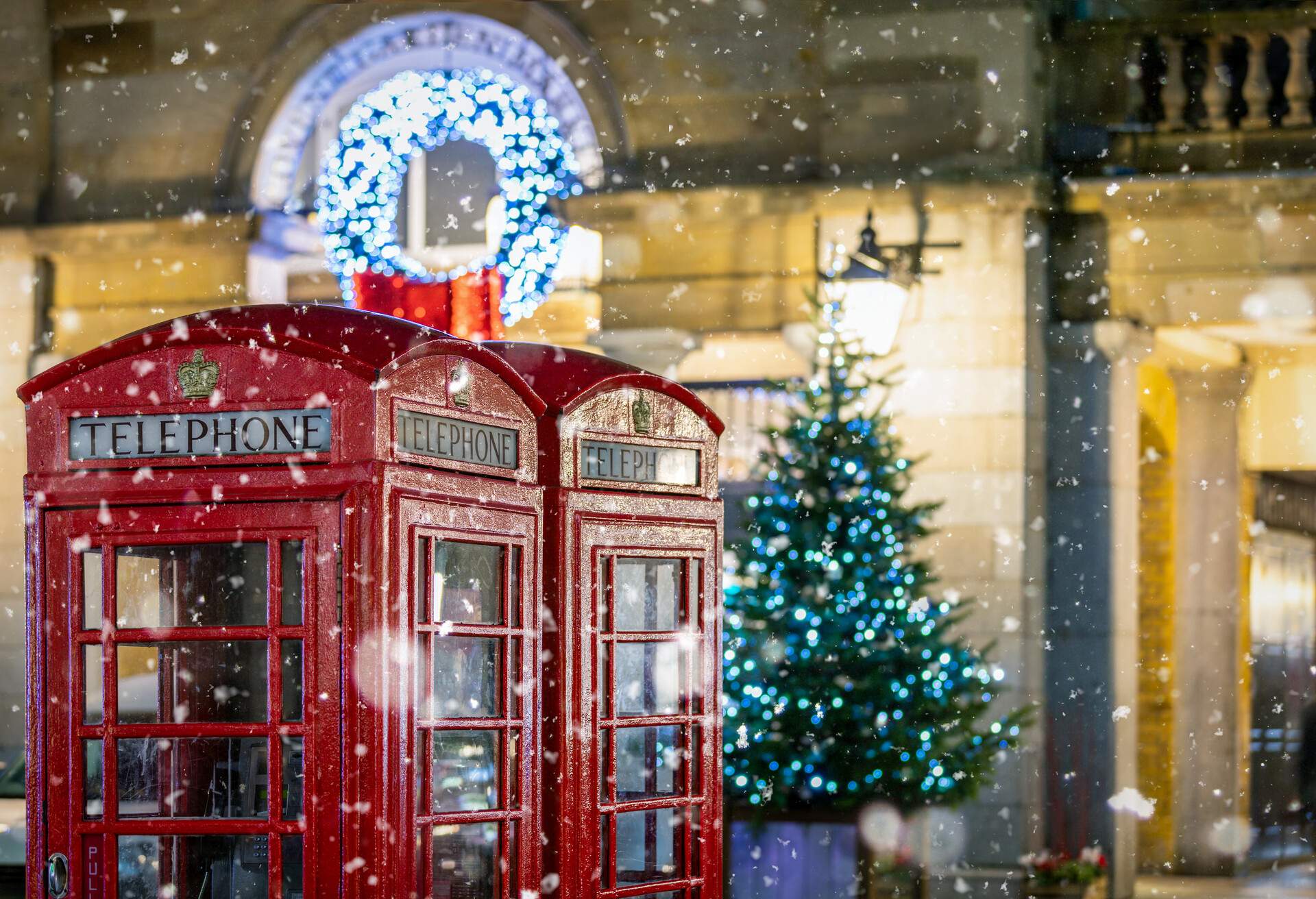 Two iconic red London telephone booths and an illuminated Christmas tree in the back, on a snowy winter night.