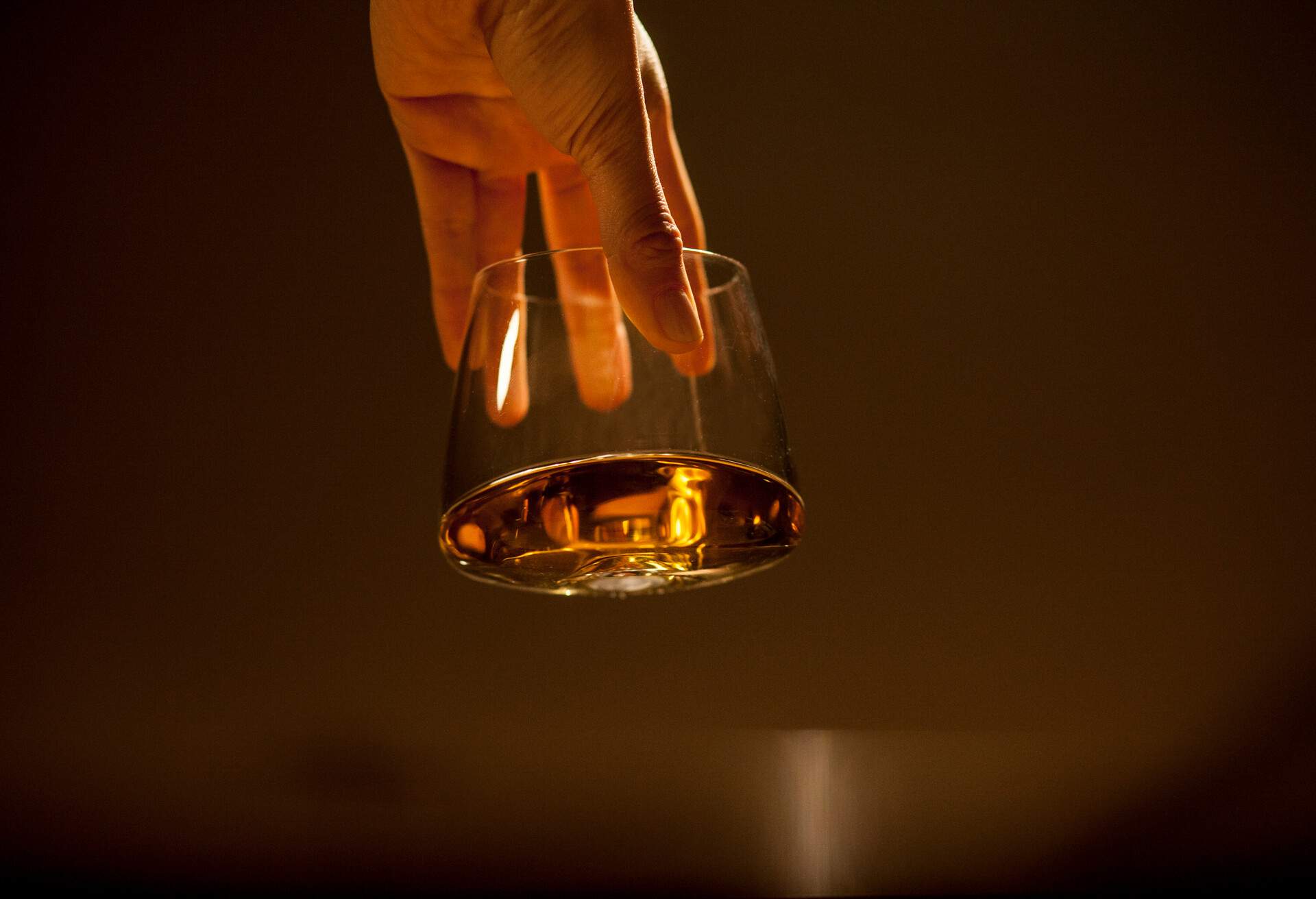 Portrait of a hand holding a transparent glass with golden liquor on a brownish background.