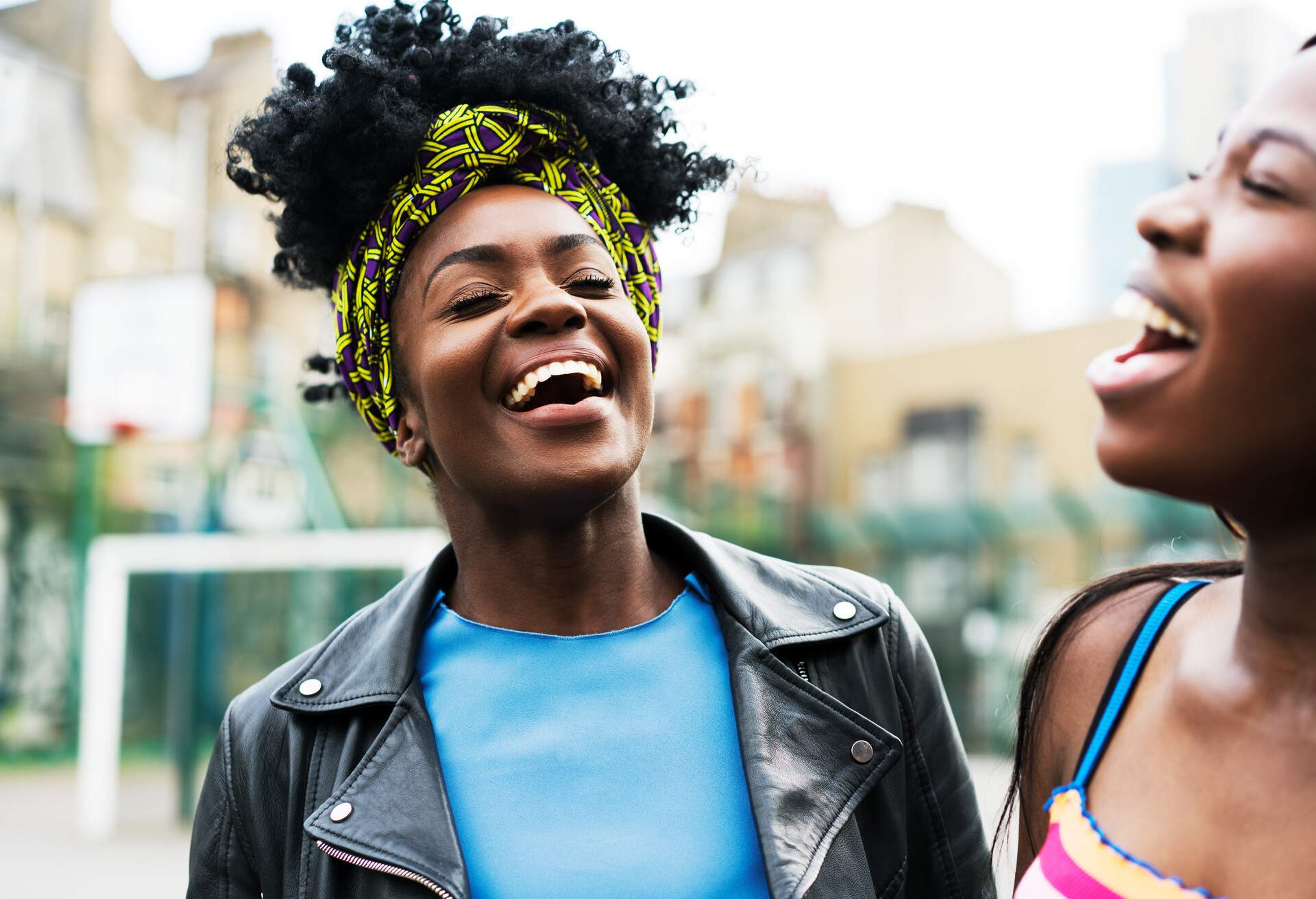 A curly-haired woman in a colourful bandana and black leather jacket laughs with a friend.