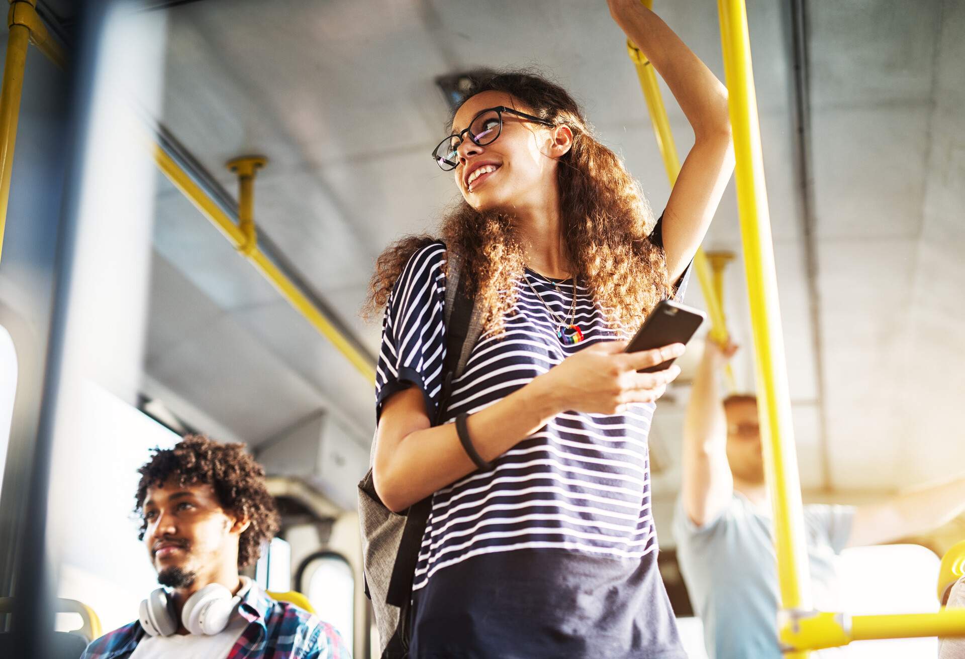 A young woman standing on a bus, holding her phone and glancing behind her.