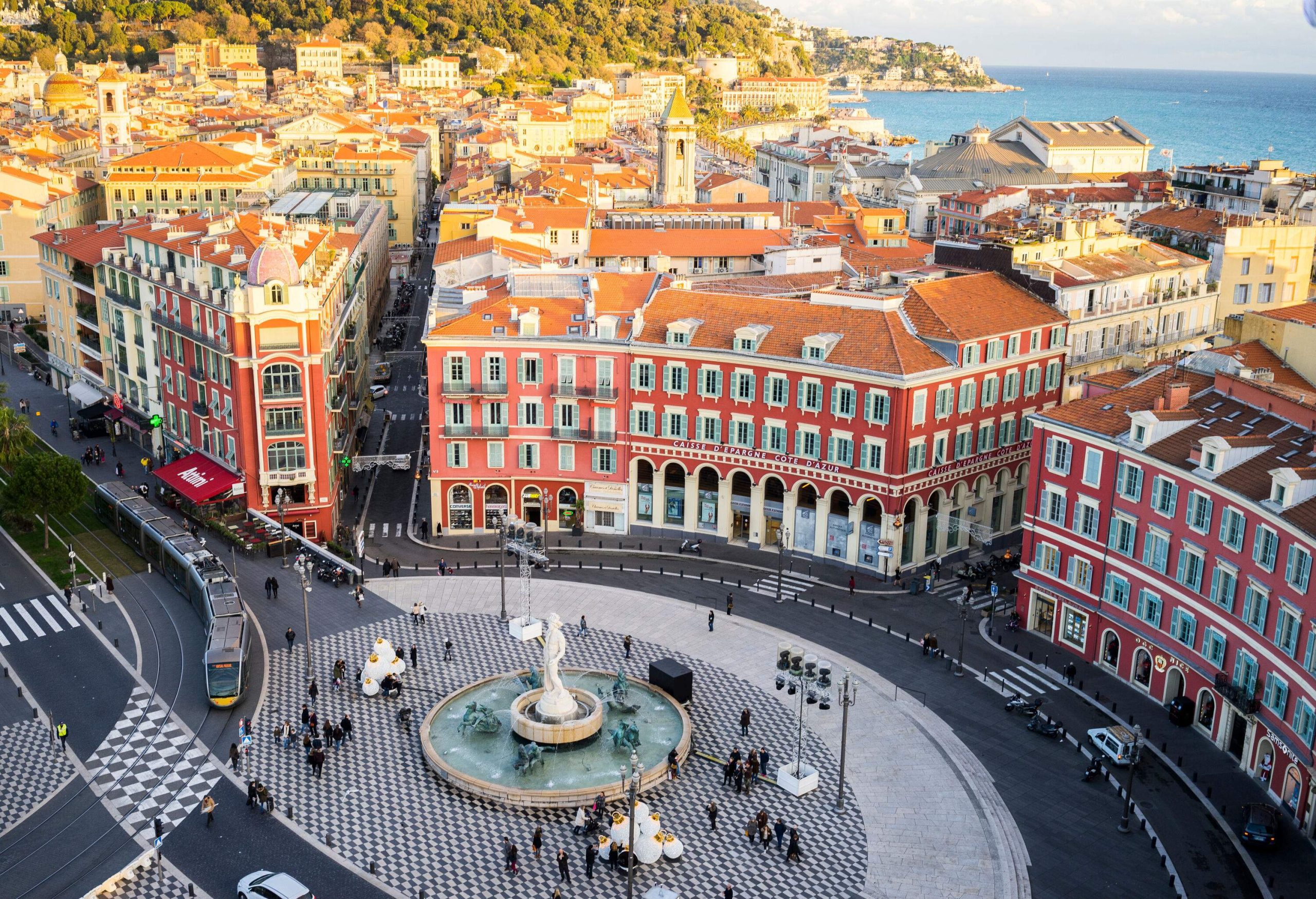 A circular fountain with a statue in an open square surrounded by red-coloured buildings and a distant view of the sea.