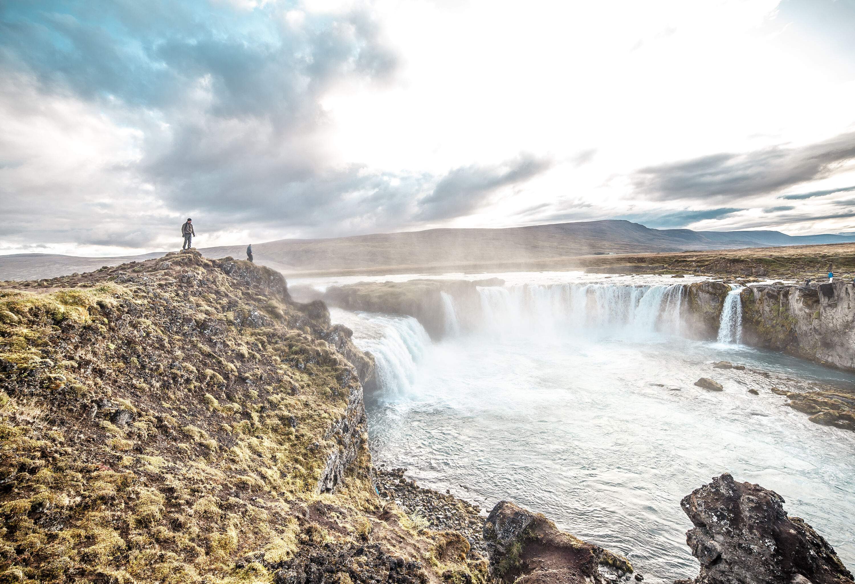 A man stands at the edge of a rock cliff with views of a waterfall that pours into a raging river.