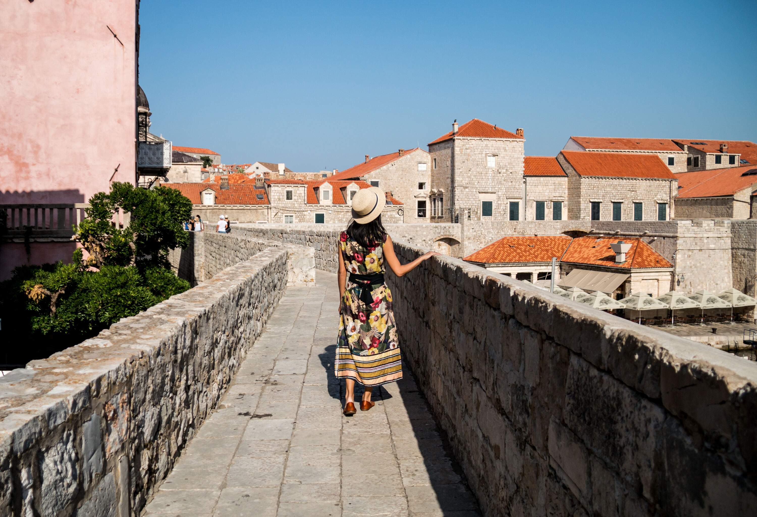 A woman leisurely strolls along the historic Walls of Dubrovnik, with stone buildings as a picturesque backdrop.