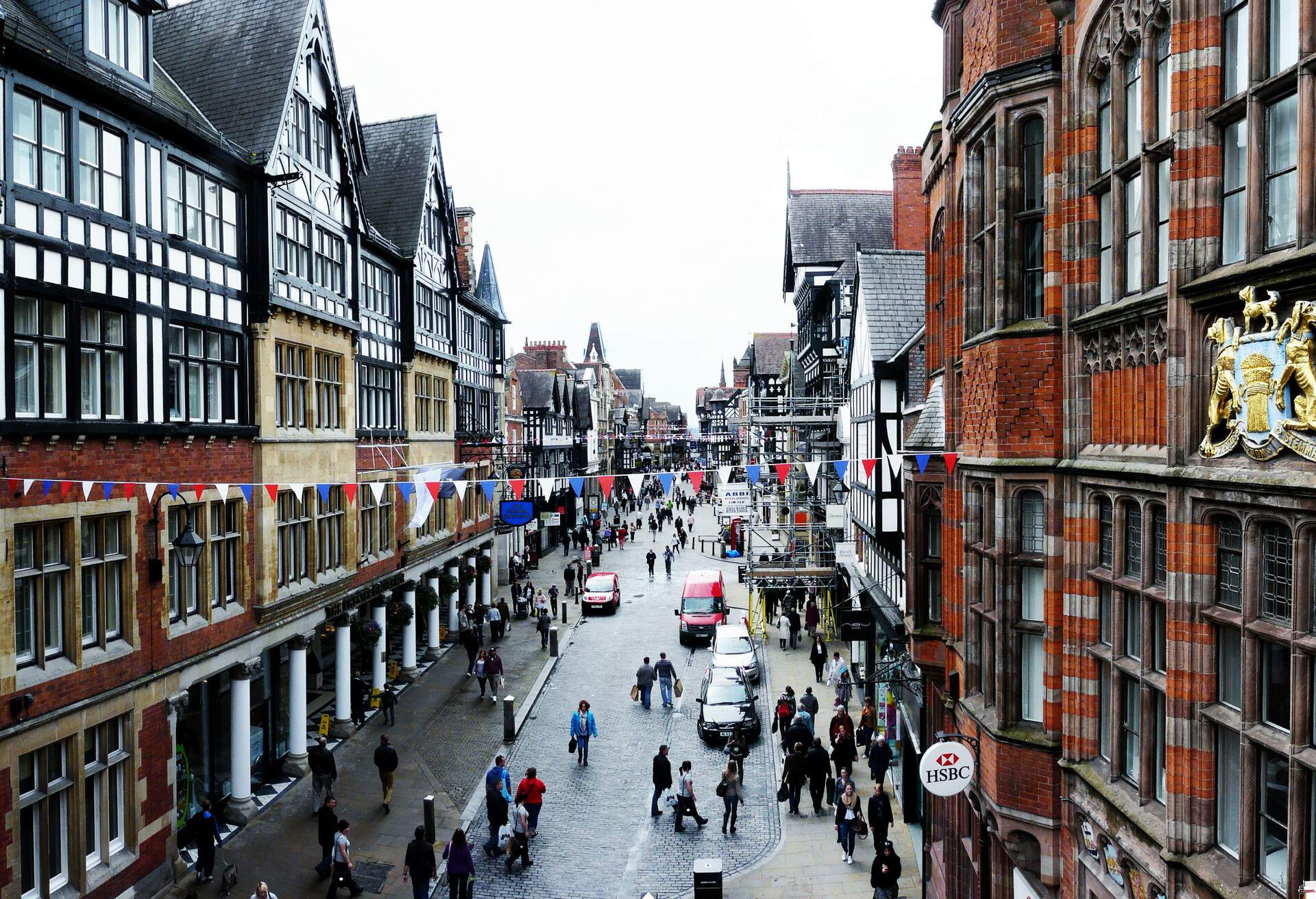 A crowded cobbled street lined on both sides by tall buildings in an overcast.