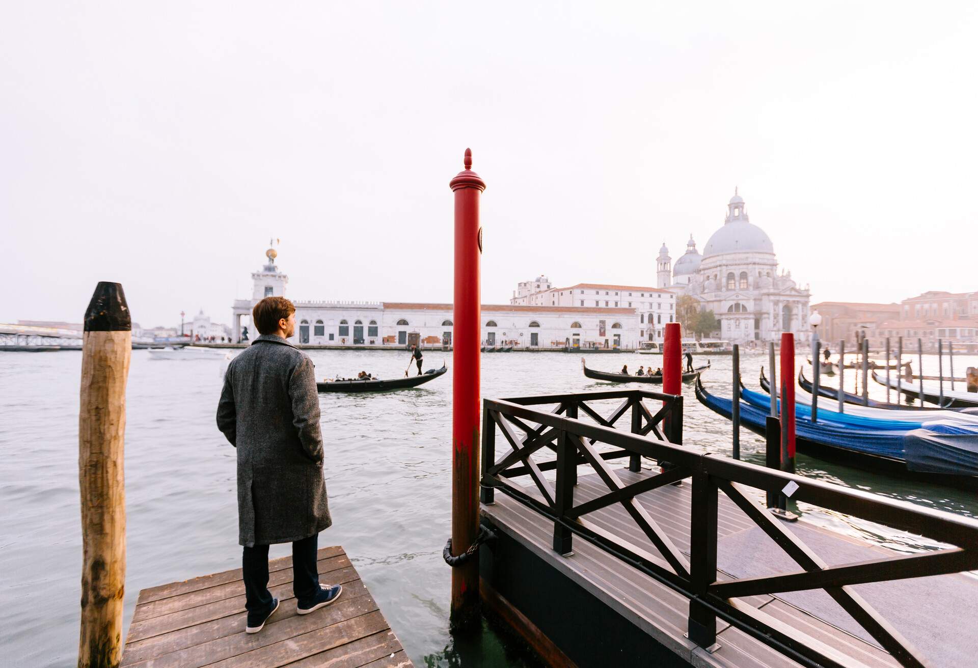 A person donning a long coat stands pensively on the pier, gazing intently at a majestic white building adorned with a grand dome across the shimmering waters.