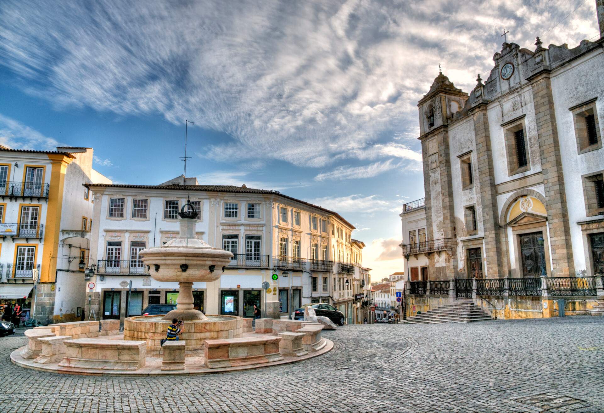 A circular marble fountain in the middle of a cobbled square confined with buildings.