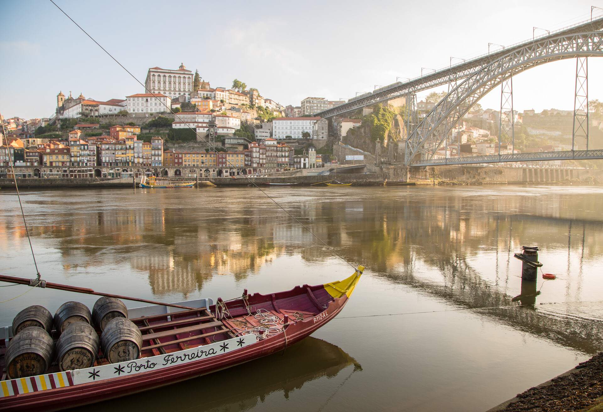 A laden ship carrying barrels navigates the river, passing under a bridge while the picturesque waterfront buildings stand proudly across the water.