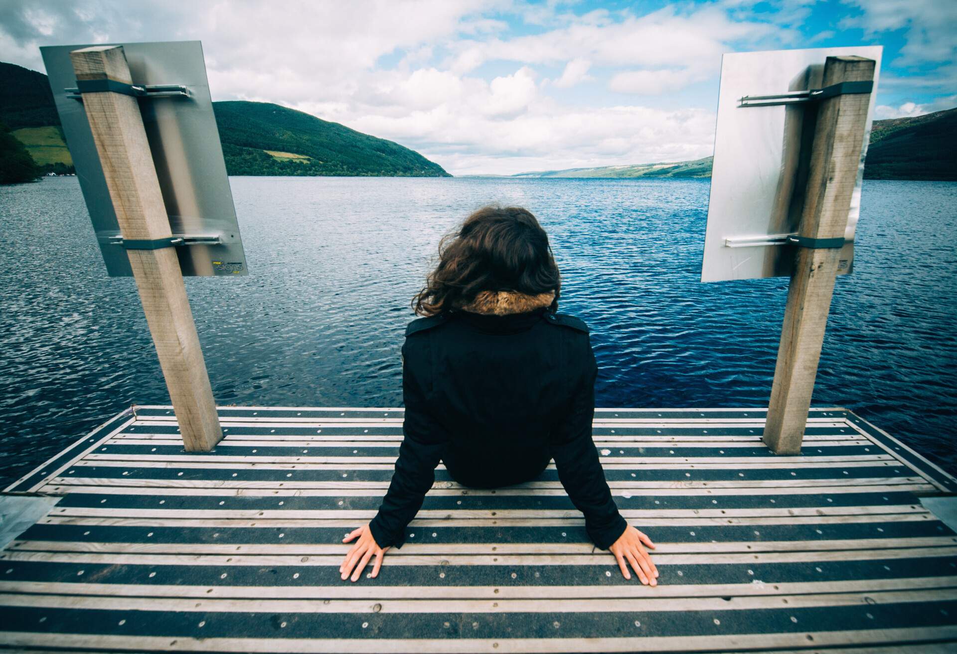 A young woman sits on a dock with a view of a  large freshwater lake surrounded by lush mountains.