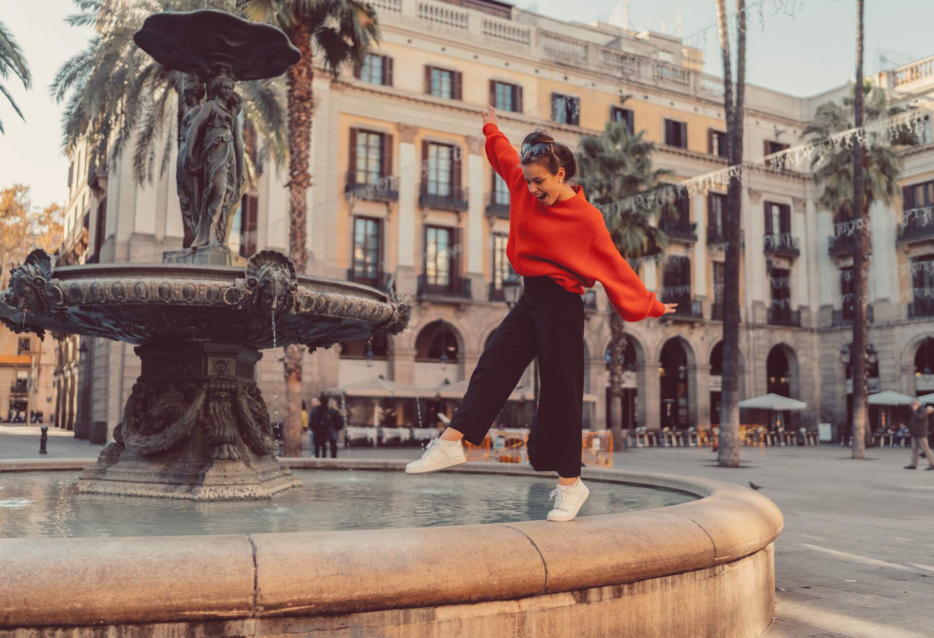 A happy lady balancing at the edge of a fountain in the city square enclosed by buildings.