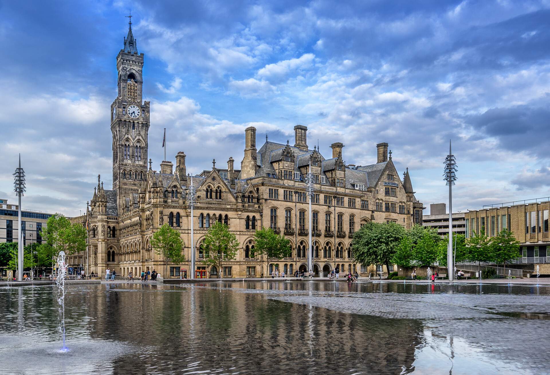 A Gothic-style town hall and clock tower in a square with a ground water fountain.