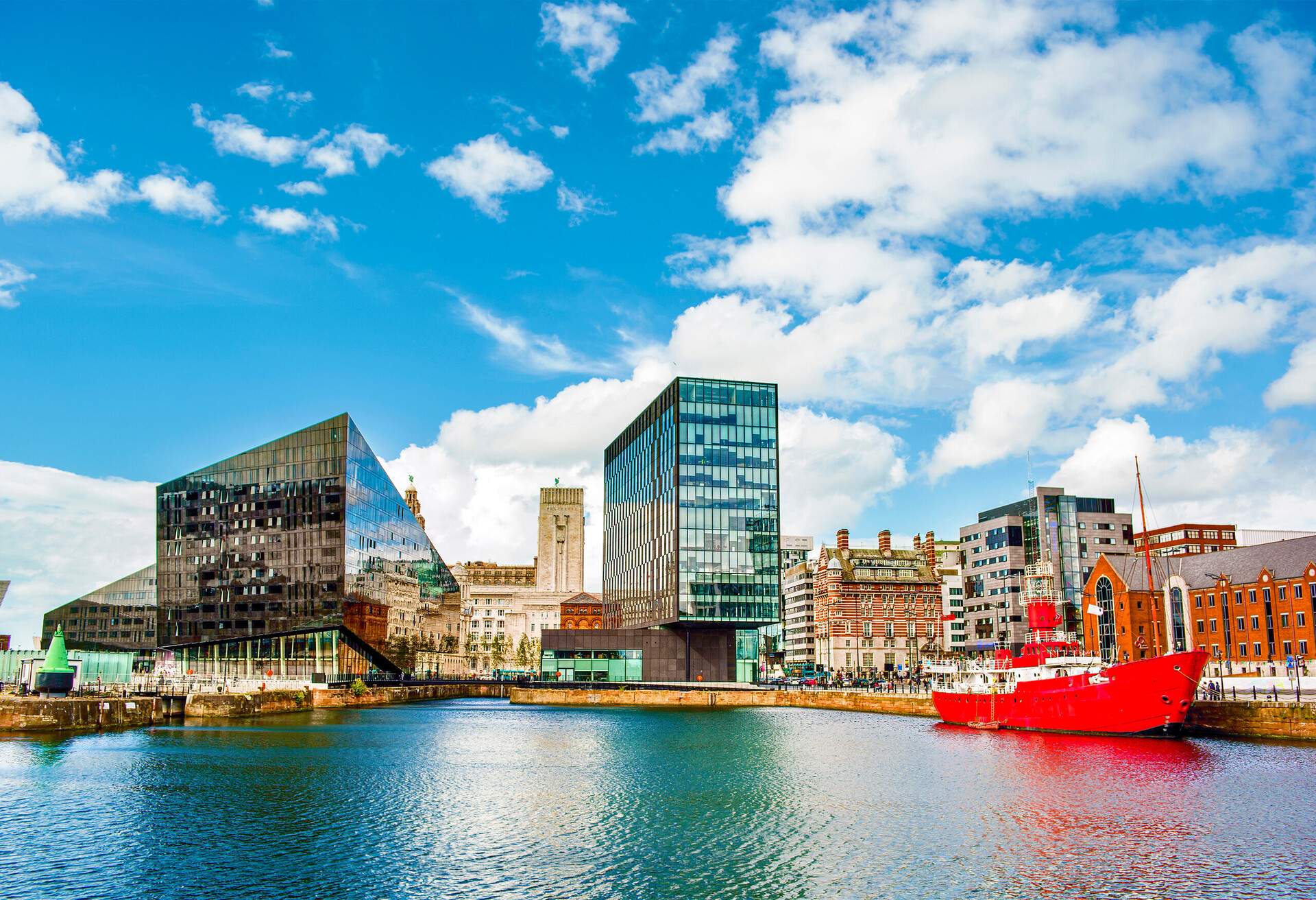 A small red ship docked along a river lined with colourful buildings near a picturesque two adjacent glass buildings under a cloudy blue sky.