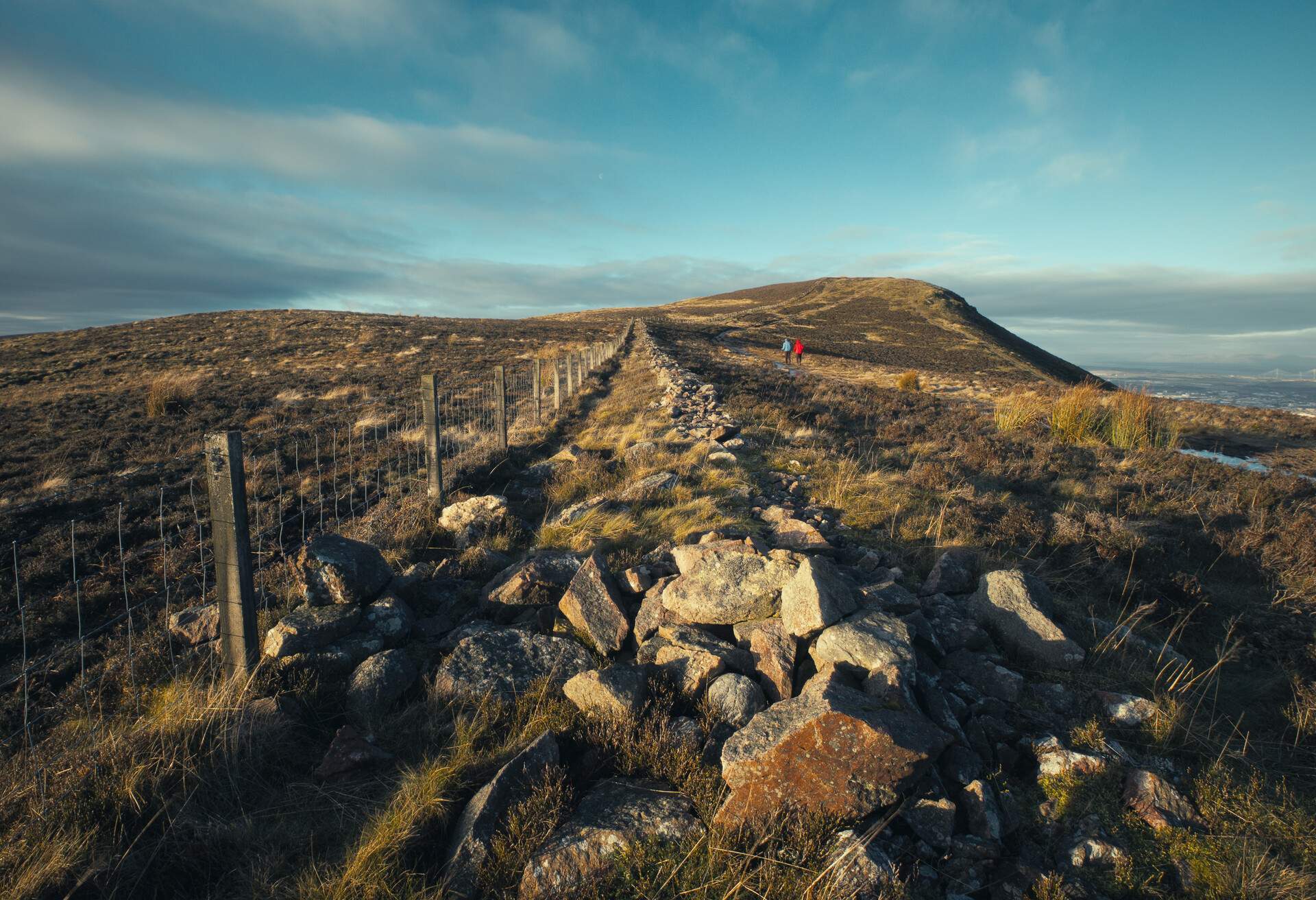 Hiking in Pentland Hills Regional Park. In the foreground are stones and a fence stretching into the distance. In the background, people from the back hiking in the hills