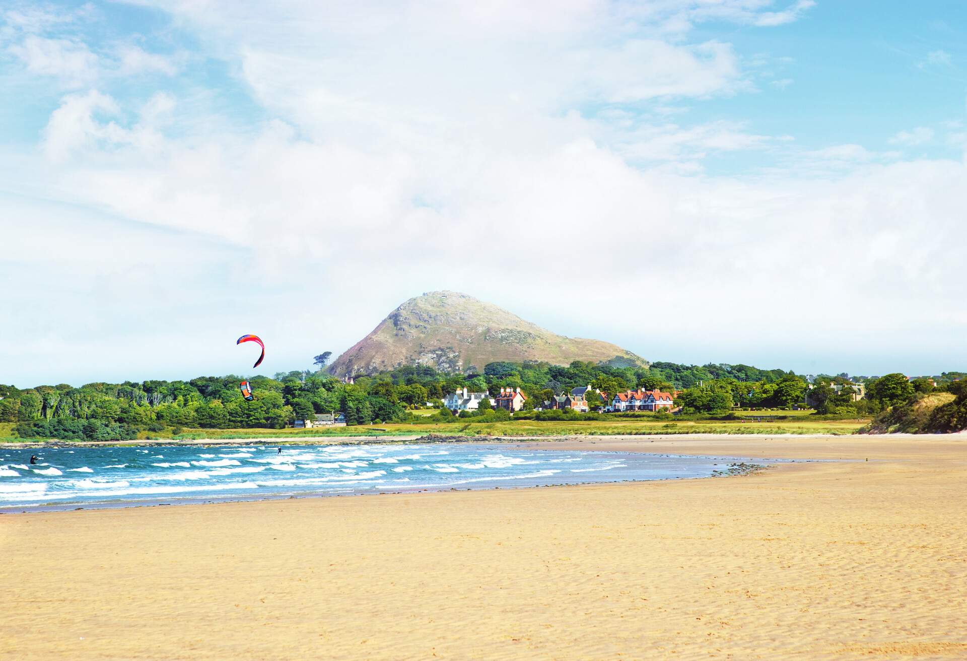 Windsurfers riding the turbulent waves along a broad beach lined with residences and tree groves.