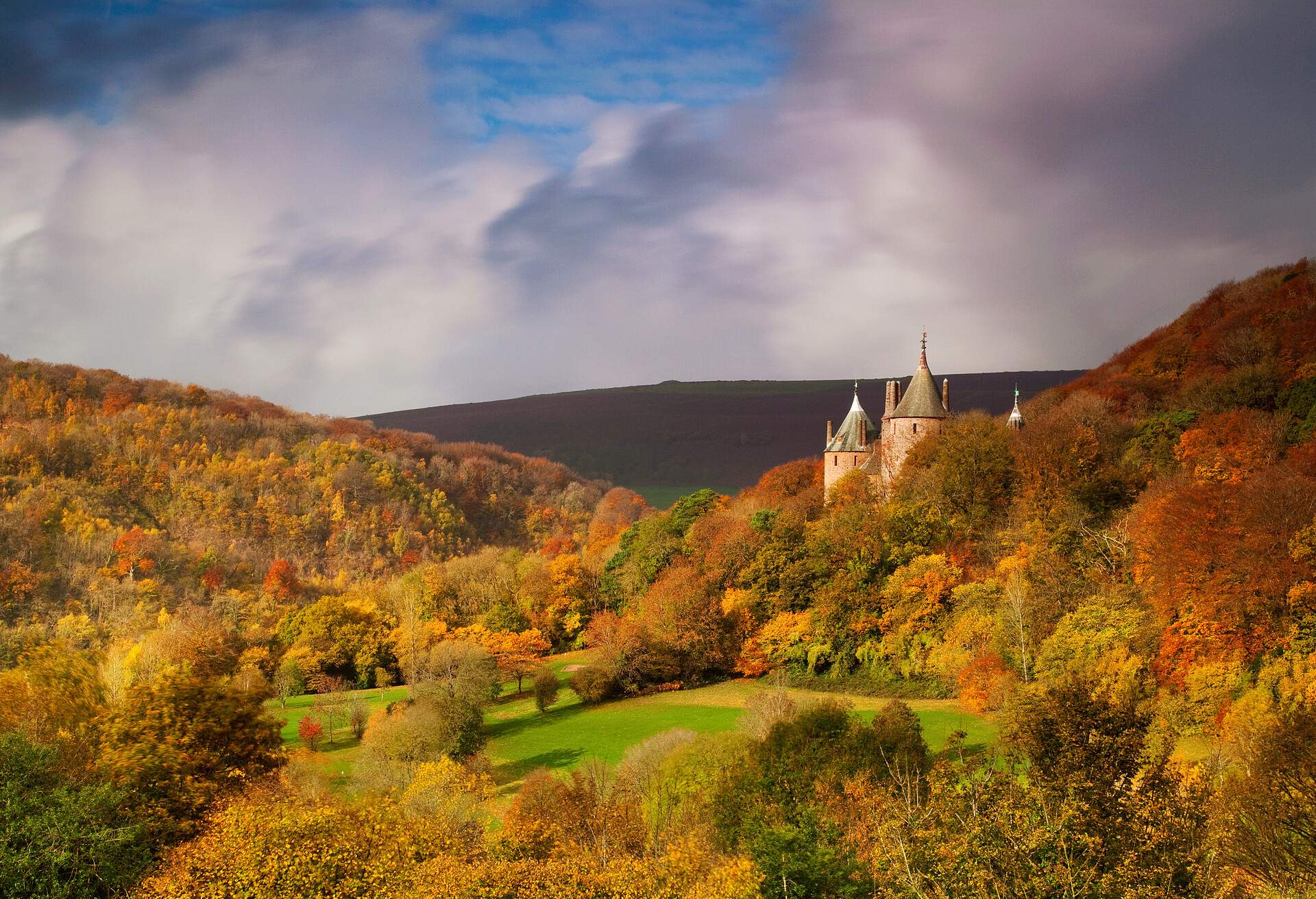 A castle lies on a mountainside covered with lush trees with a view of a flat mountaintop in the background in autumn.