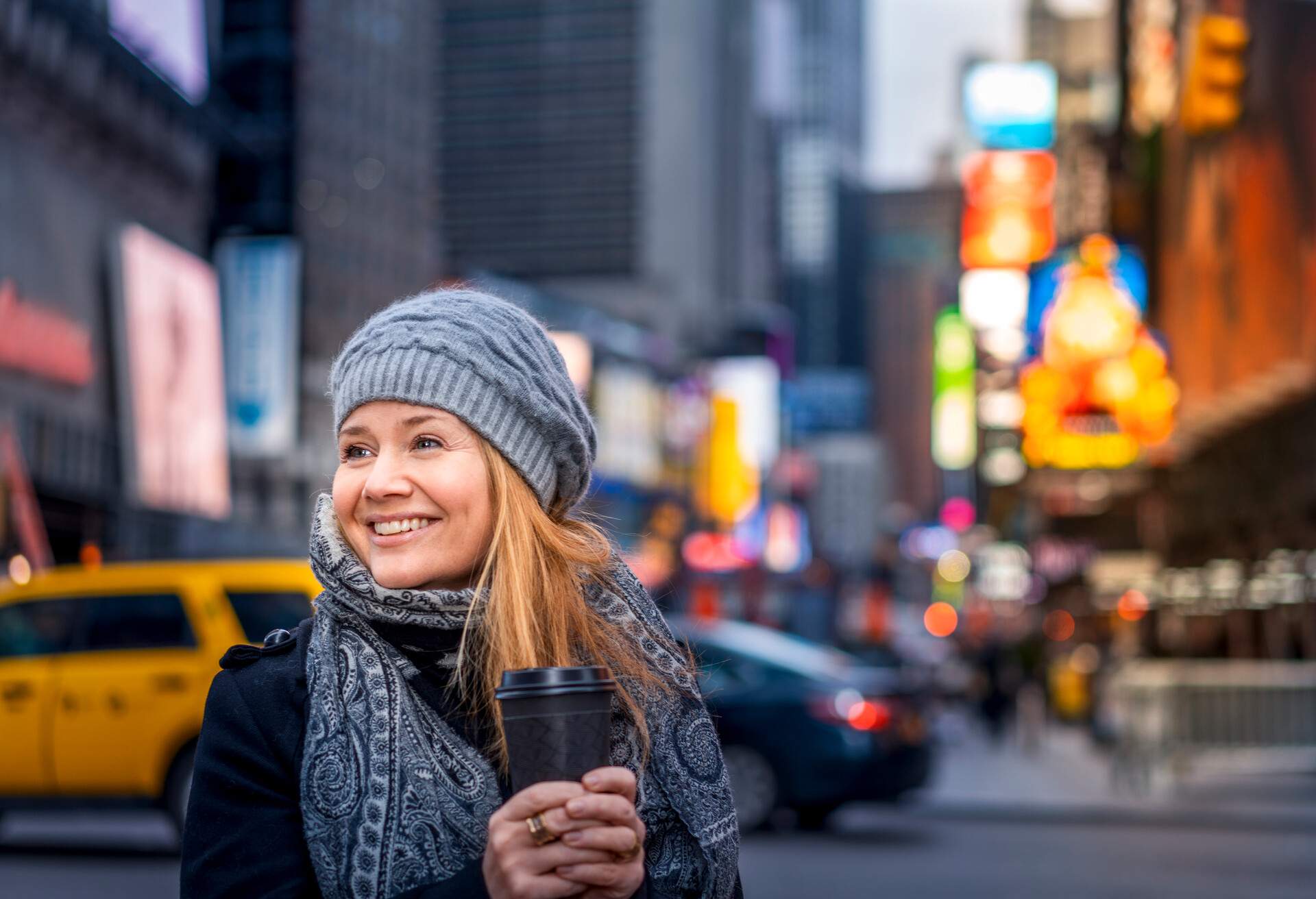 A woman in a grey bonnet and printed scarf smiles as she holds a cup of coffee.