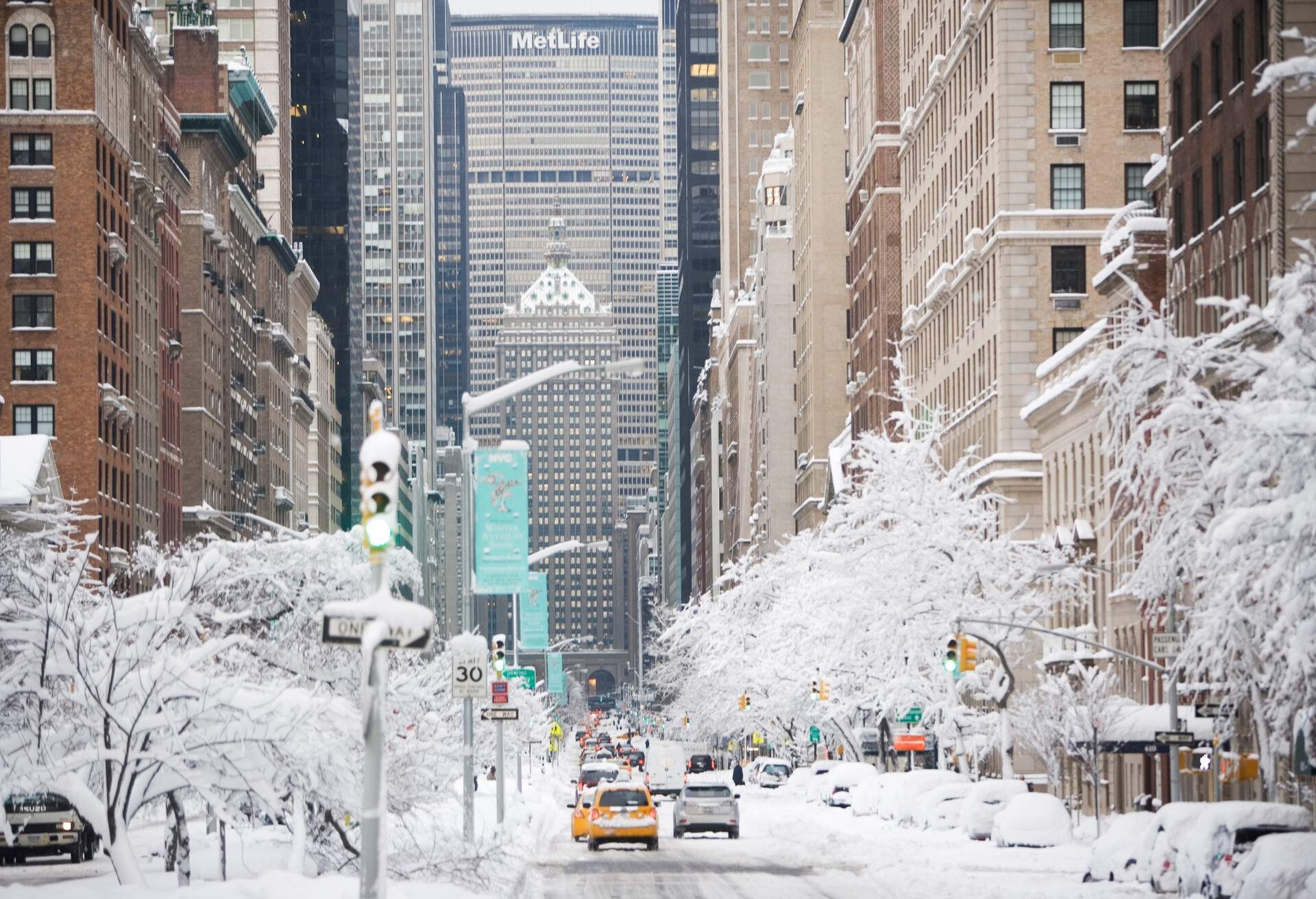 Cars travelling on a snowy highway bordered by frozen trees and adjacent high-rise buildings.