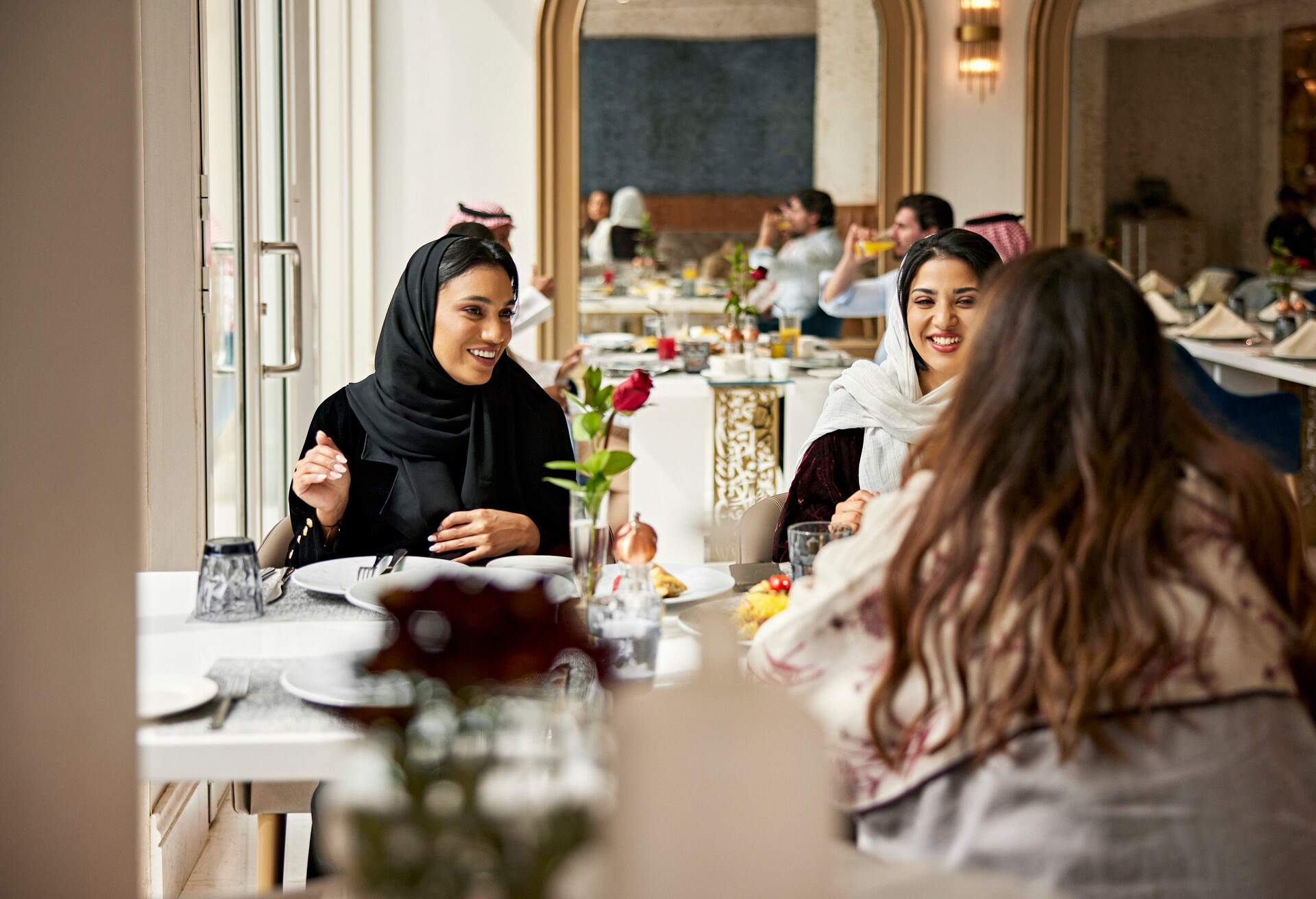 A group of women wearing scarves dining at a restaurant while cheerfully chatting with one another.