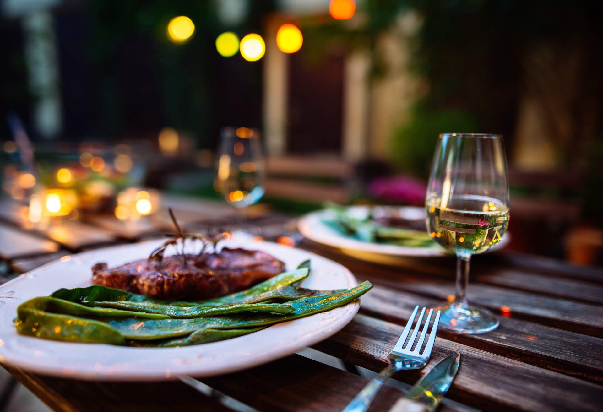 A romantic outdoor dinner for two, featuring a wooden table adorned with elegant tableware, a deliciously cooked steak on a plate, and wine glasses, accompanied by a set of silverware, is ready for a delightful dining experience.
