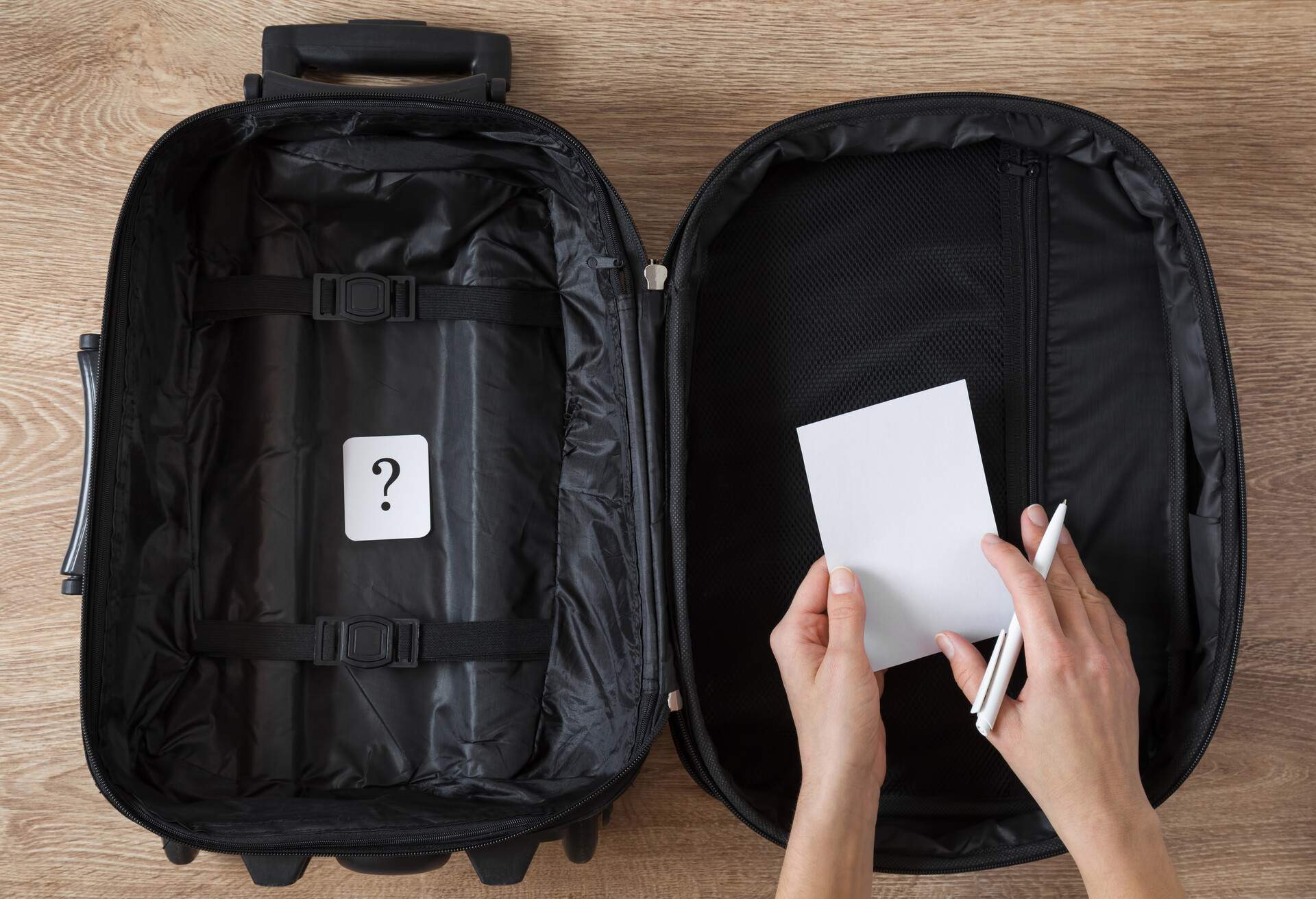 An opened empty, black travel bag with a question mark written on a white card and an individual holding a piece of paper and a pen.