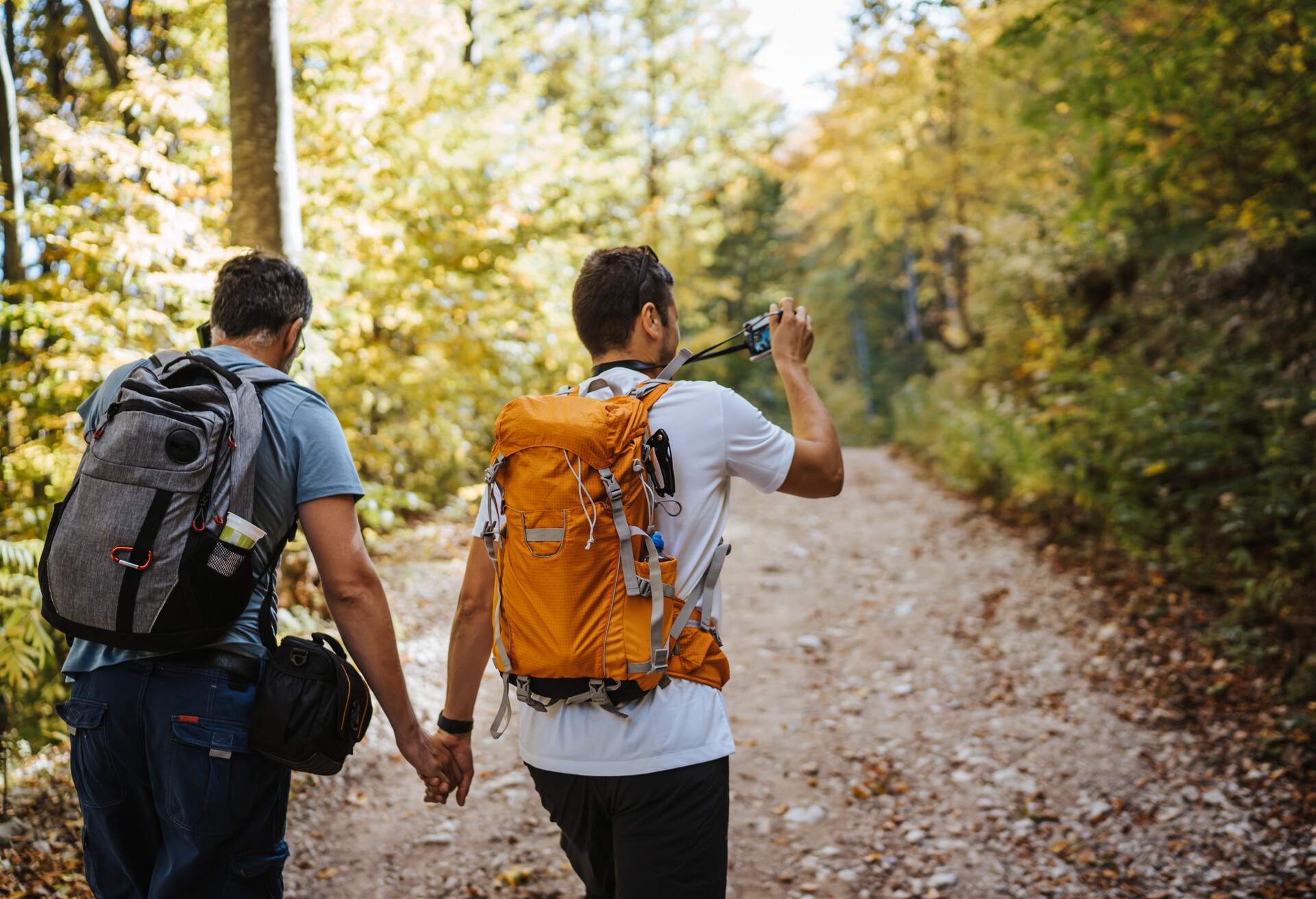 Male backpackers hiking in the woods while holding hands, one holding a camera and taking photos.