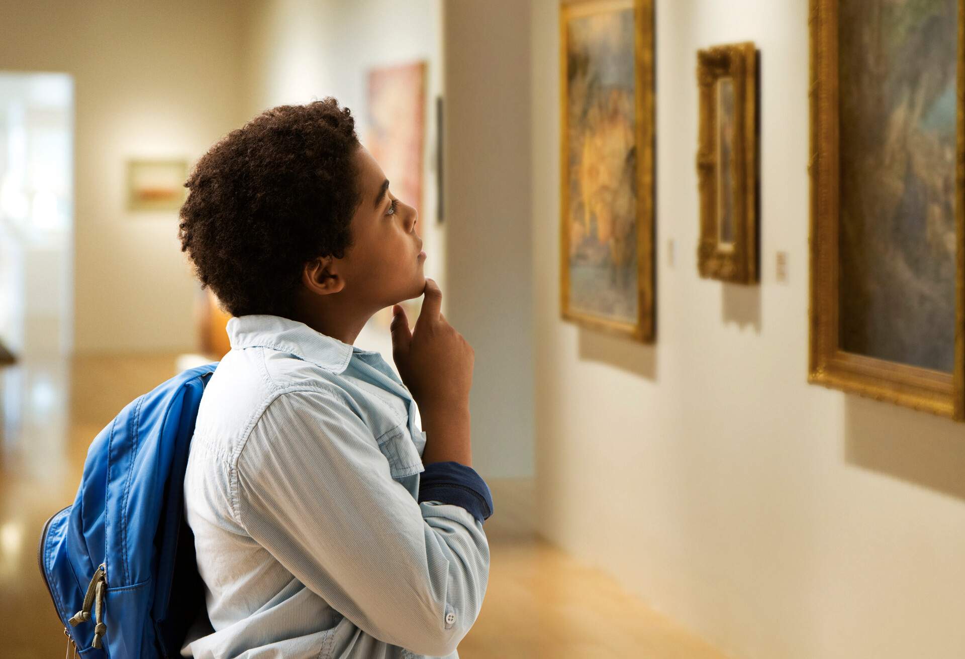 A person with a backpack gazes at a painting, hands thoughtfully resting on their chin, as they appear to be contemplating the artwork's meaning.