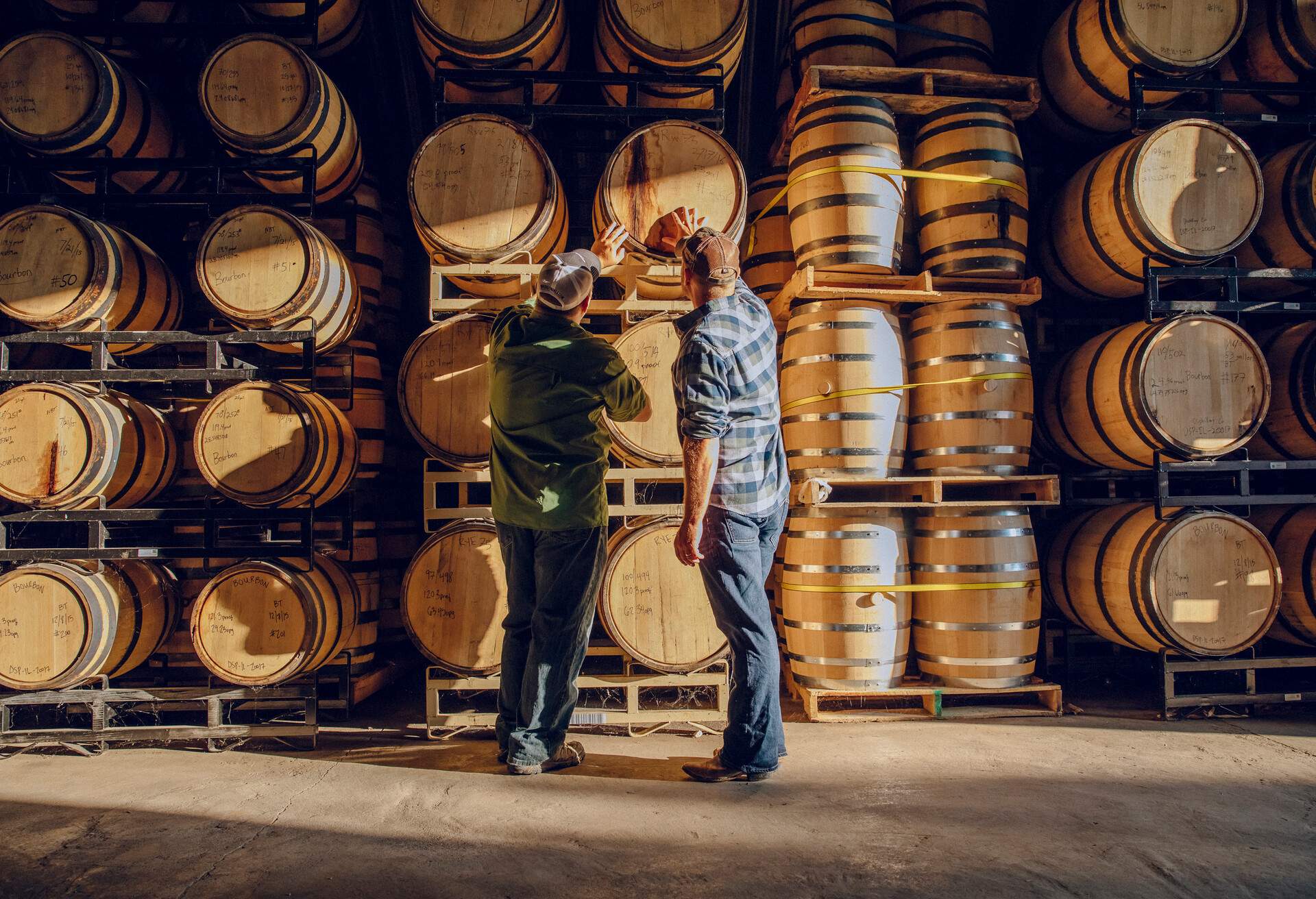 Two men looking at wooden barrels inside a distillery.
