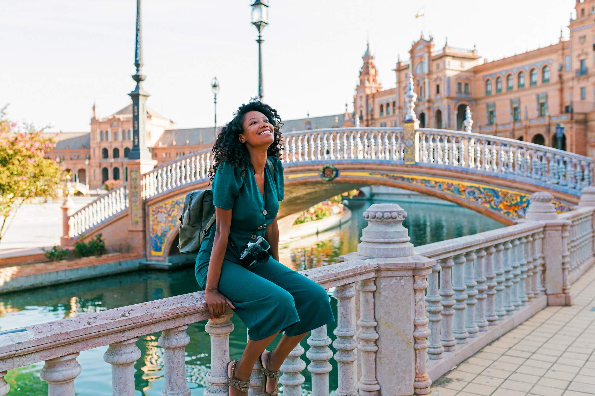 A woman in casual attire smiles as she sits on the railings of the sidewalk next to a bridge.