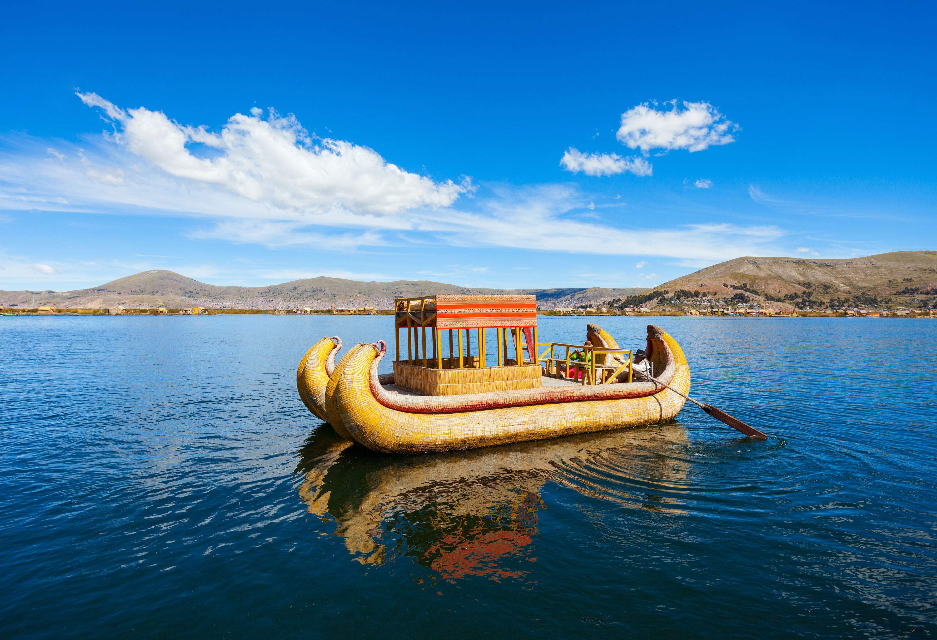 Two people ride a yellow totora boat with a square shed and use a paddle to navigate the lake.