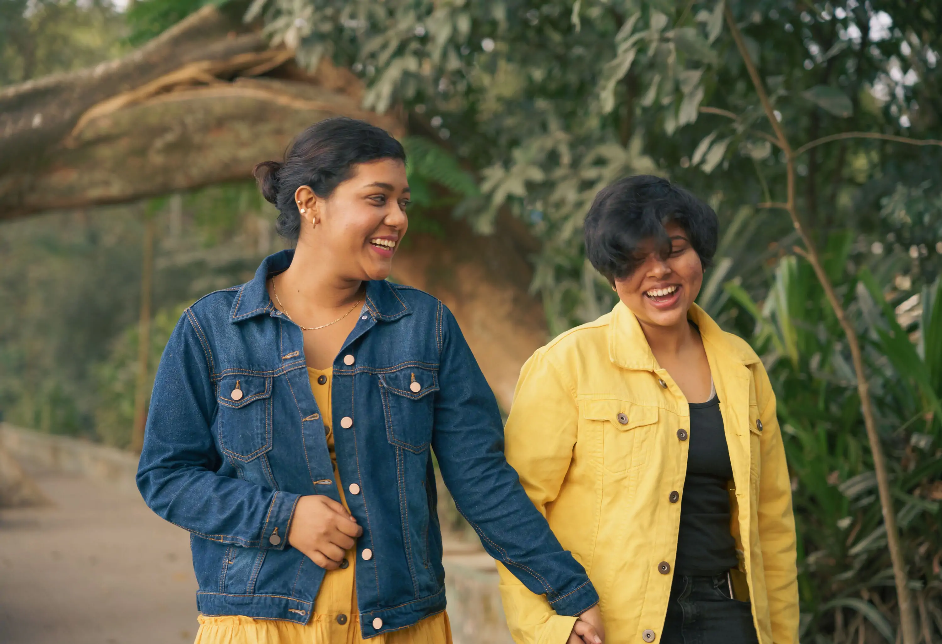 Two female friends in blue and yellow jacket hold hands while walking on the street.
