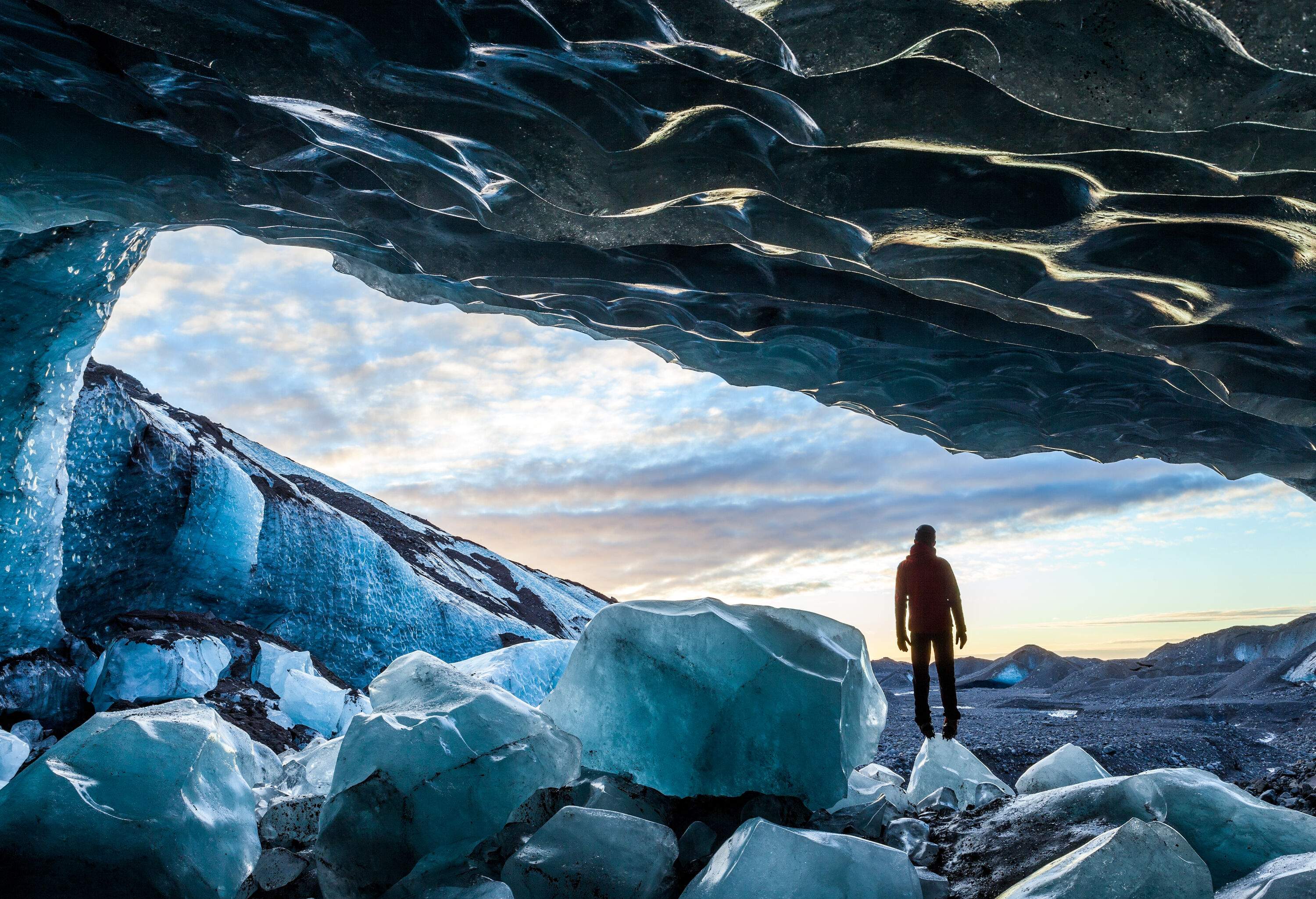 A man stands over a dramatic sky on a small, pointed glacier inside an ice cave.