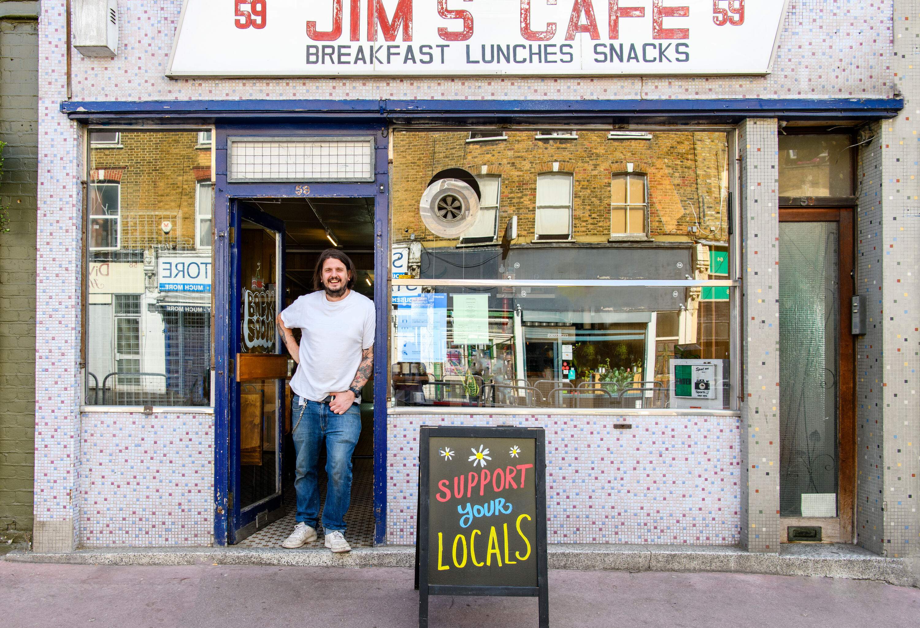 A restaurant owner smiles as he leans on the door of his restaurant with a signage in front.