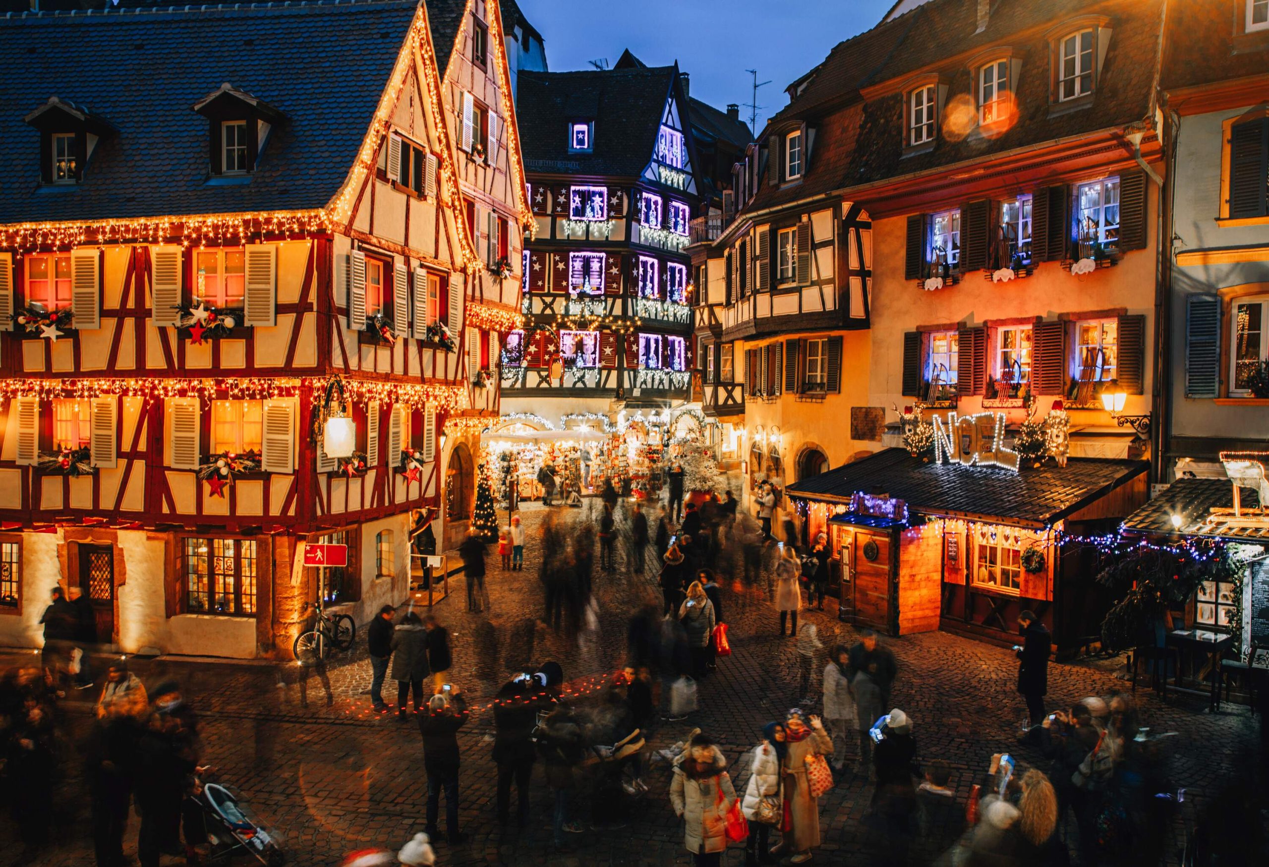 A busy street is full of crowds surrounded by half-timbered houses brightly lit with Christmas lights and decorated with various colourful Christmas ornaments.